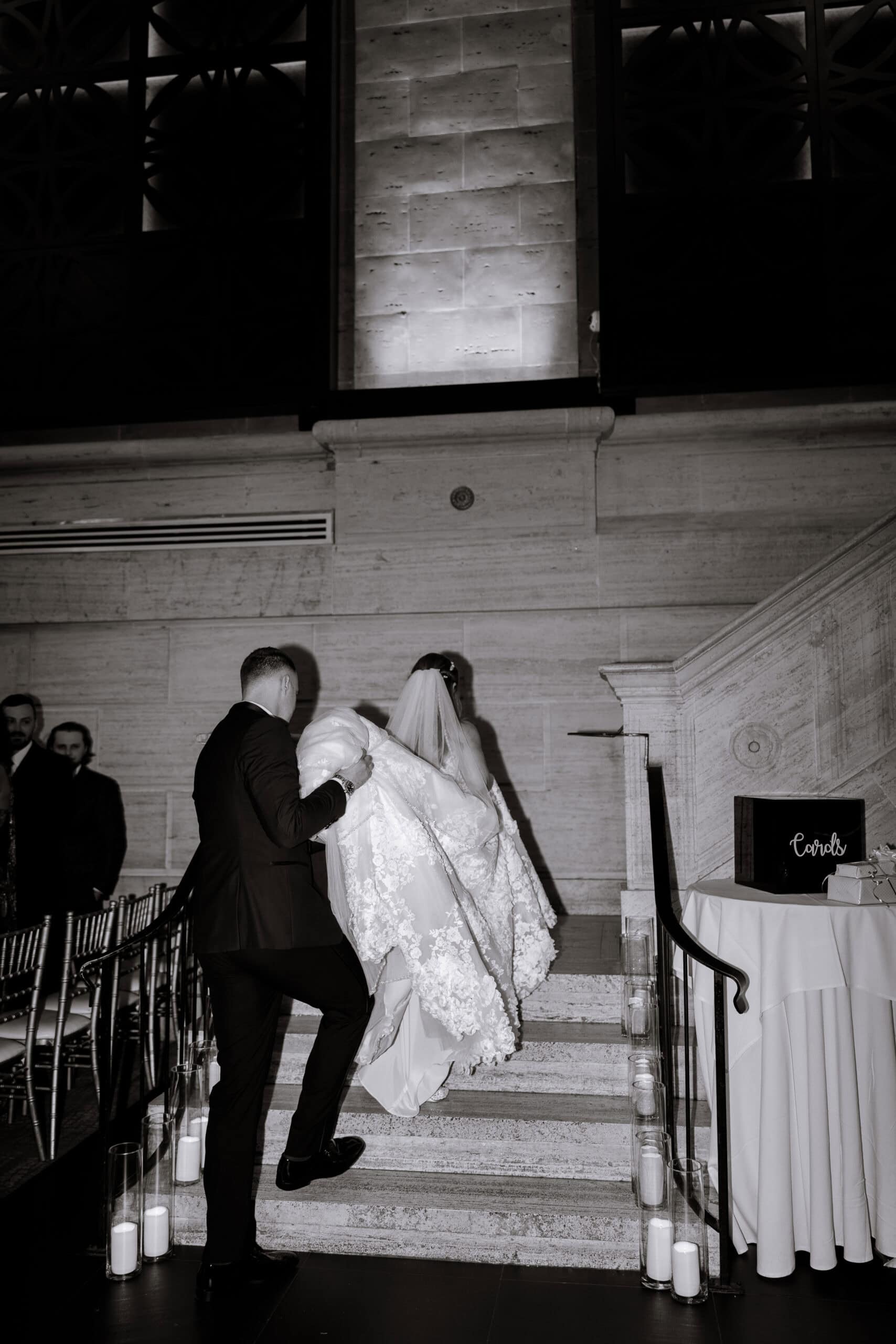 Groom carrying bride's lace wedding dress up marble stairs lined with candles at Union Trust, Philadelphia.
