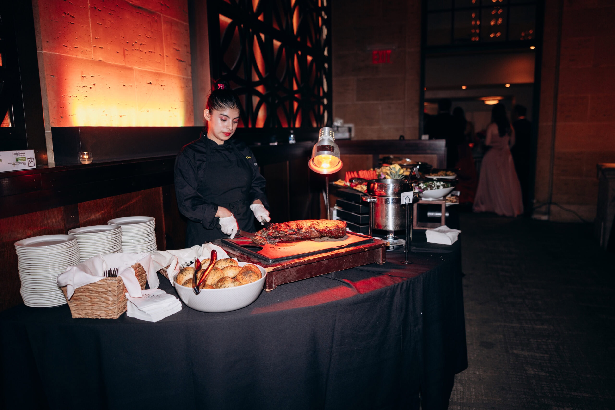 Chef in black uniform carving roasted meat at a buffet table with bread rolls and plates, warm ambient lighting, Union Trust venue