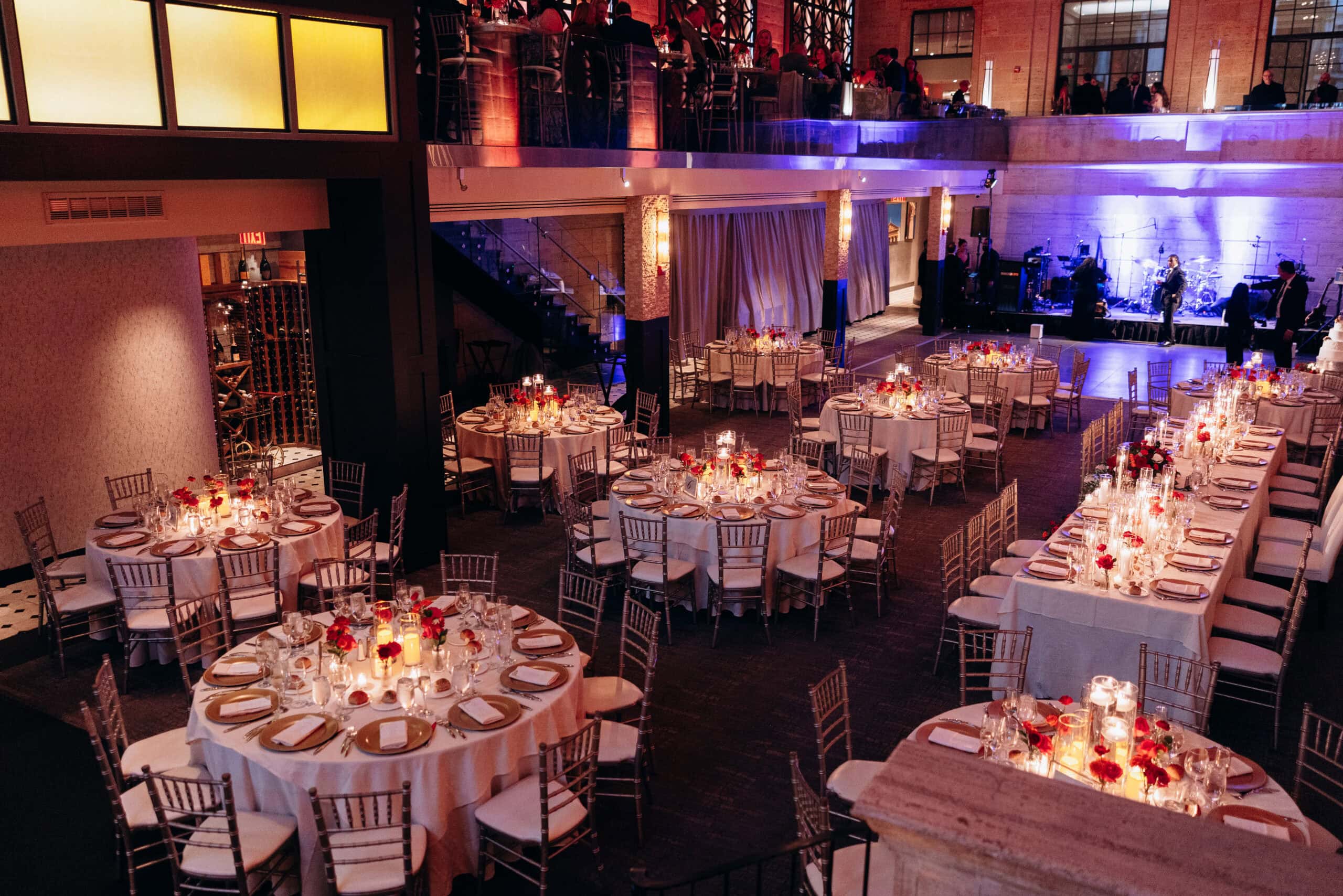 Candlelit wedding reception tables with red flowers and gold accents at Union Trust Philadelphia, with a band stage and mezzanine lounge in the background.