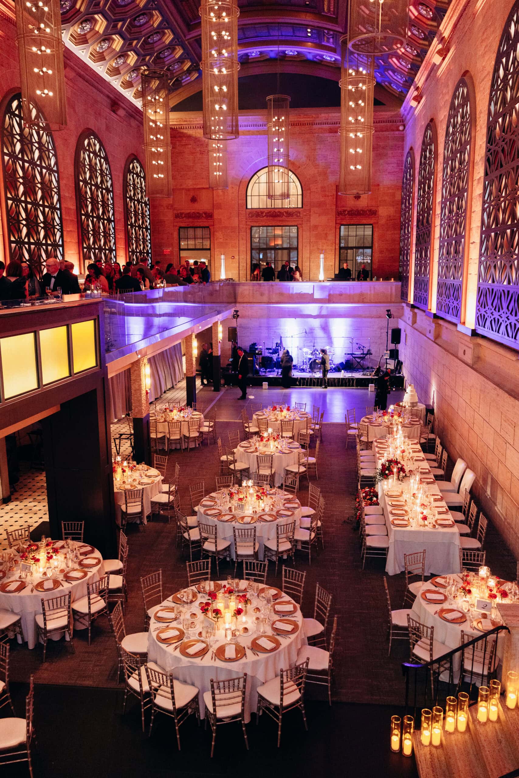 Wedding reception at Union Trust Philadelphia with round tables set with gold chargers, candles, red floral centerpieces, and a mezzanine balcony with guests.