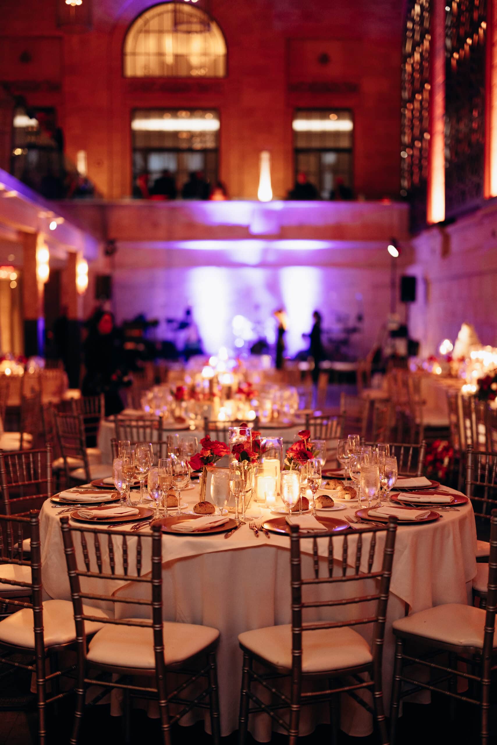 Round wedding reception table at Union Trust Philadelphia with red rose centerpieces, candles, glassware, and gold chiavari chairs in a softly lit room.