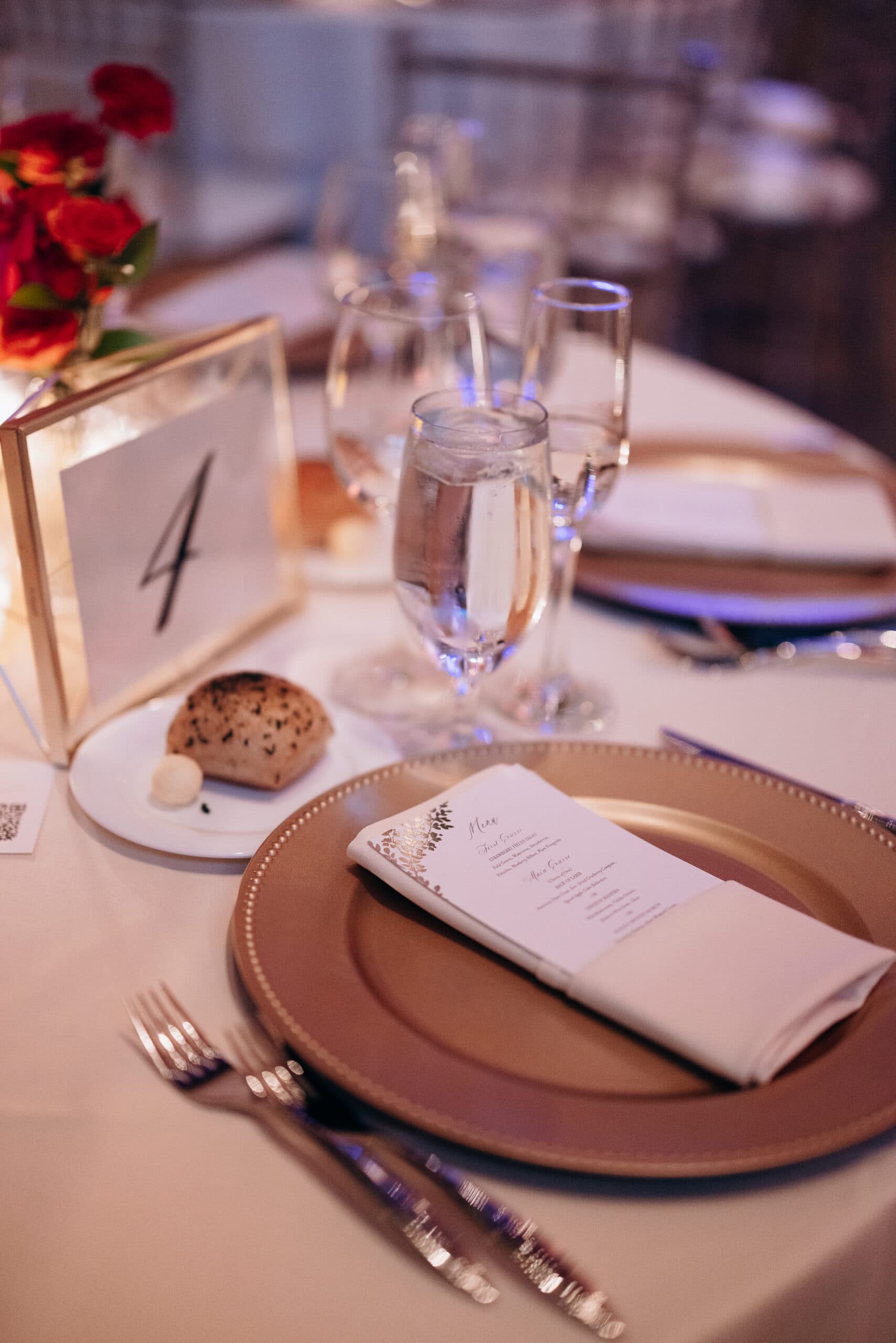 Close-up of a wedding table setting with gold charger plate, folded menu, water and champagne glasses, bread roll, and table number 4 at Union Trust, Philadelphia.