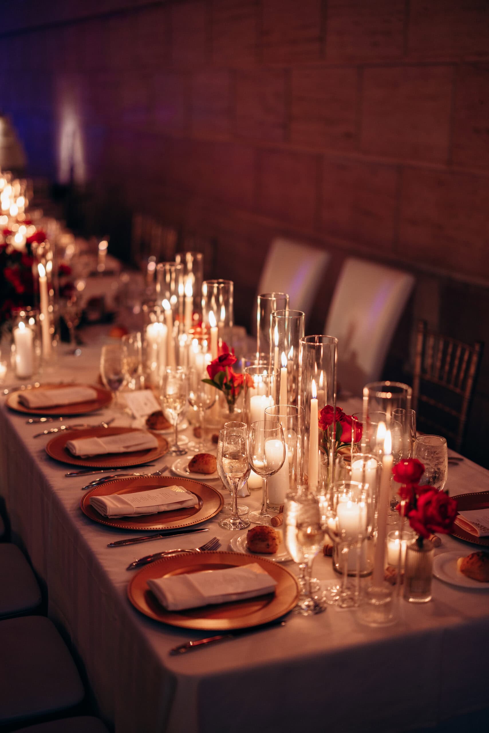 Long wedding table with white linens, gold charger plates, folded napkins, red roses, and numerous lit candles in glass holders at Union Trust, Philadelphia.
