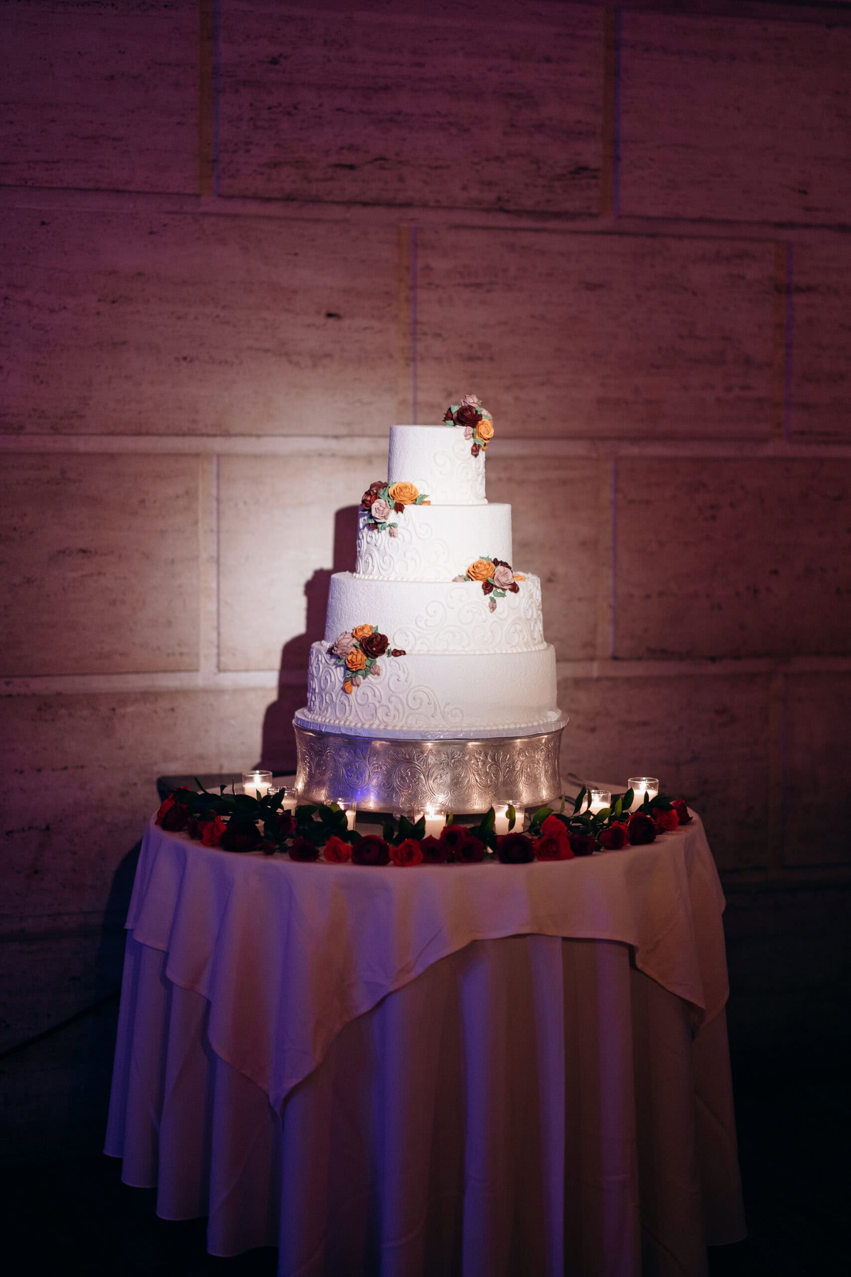 Four-tier white wedding cake with scroll detailing and orange, red, and pink floral decorations on a silver stand surrounded by candles and roses on a draped table.