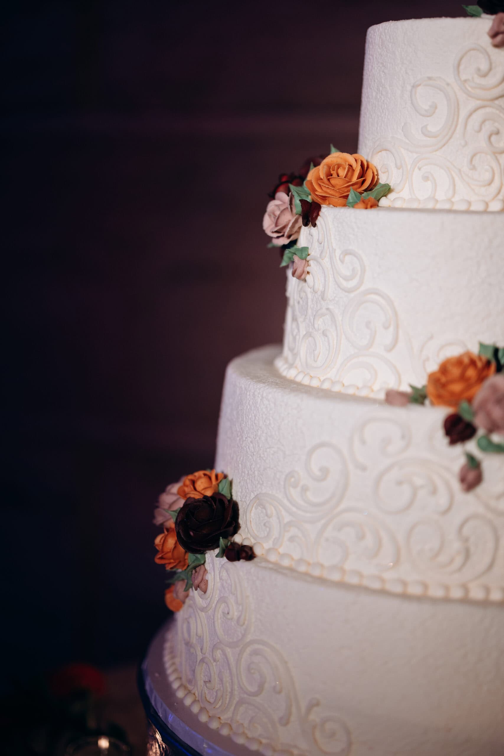 Close-up of a white tiered wedding cake with intricate scrollwork and orange, brown, and pink buttercream flowers, set against a dark background at Union Trust, Philadelphia.