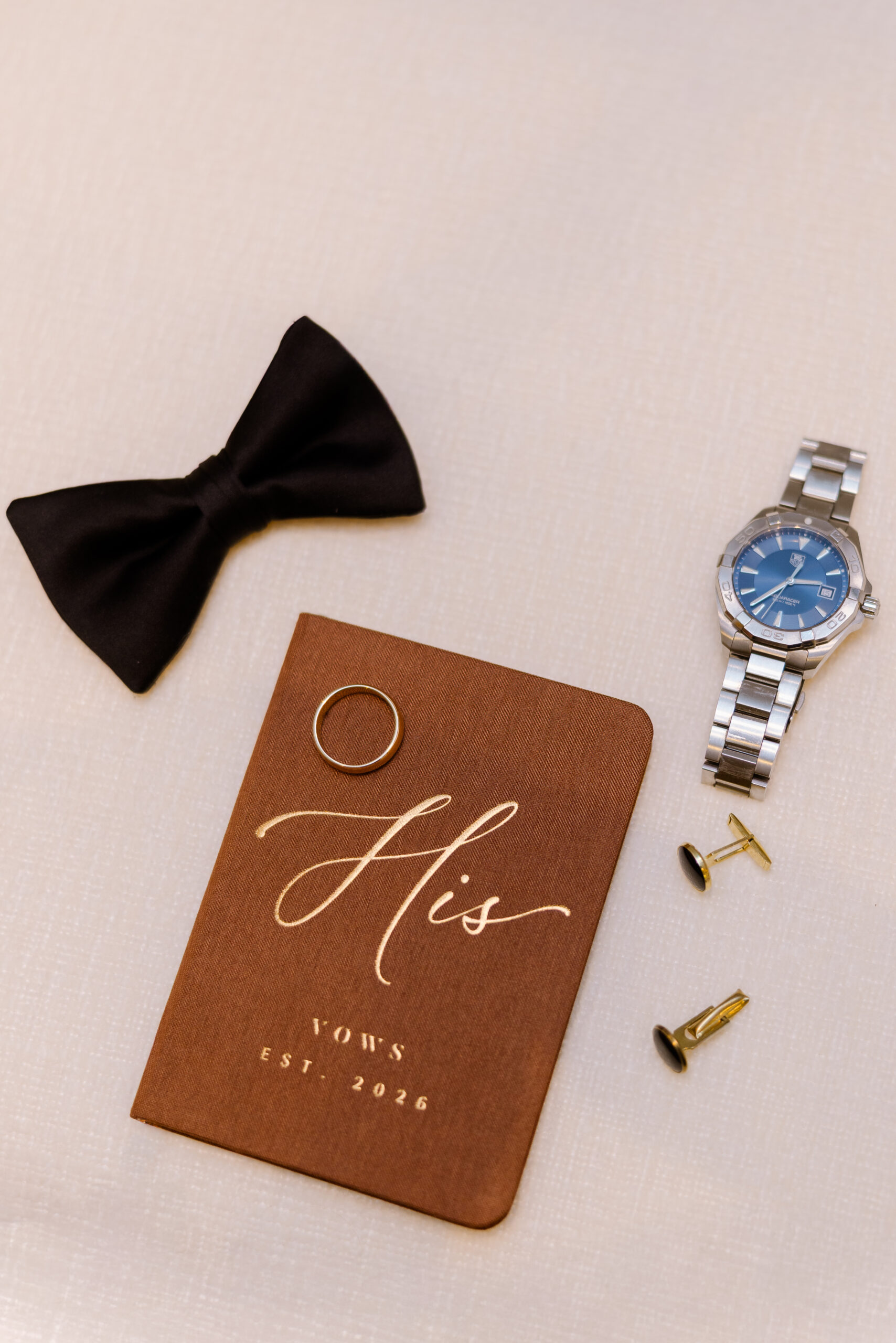 Flat lay of groom's accessories including black bow tie, silver wristwatch, gold cufflinks, and brown vow book titled 'His Vows Est. 2026' on light fabric.