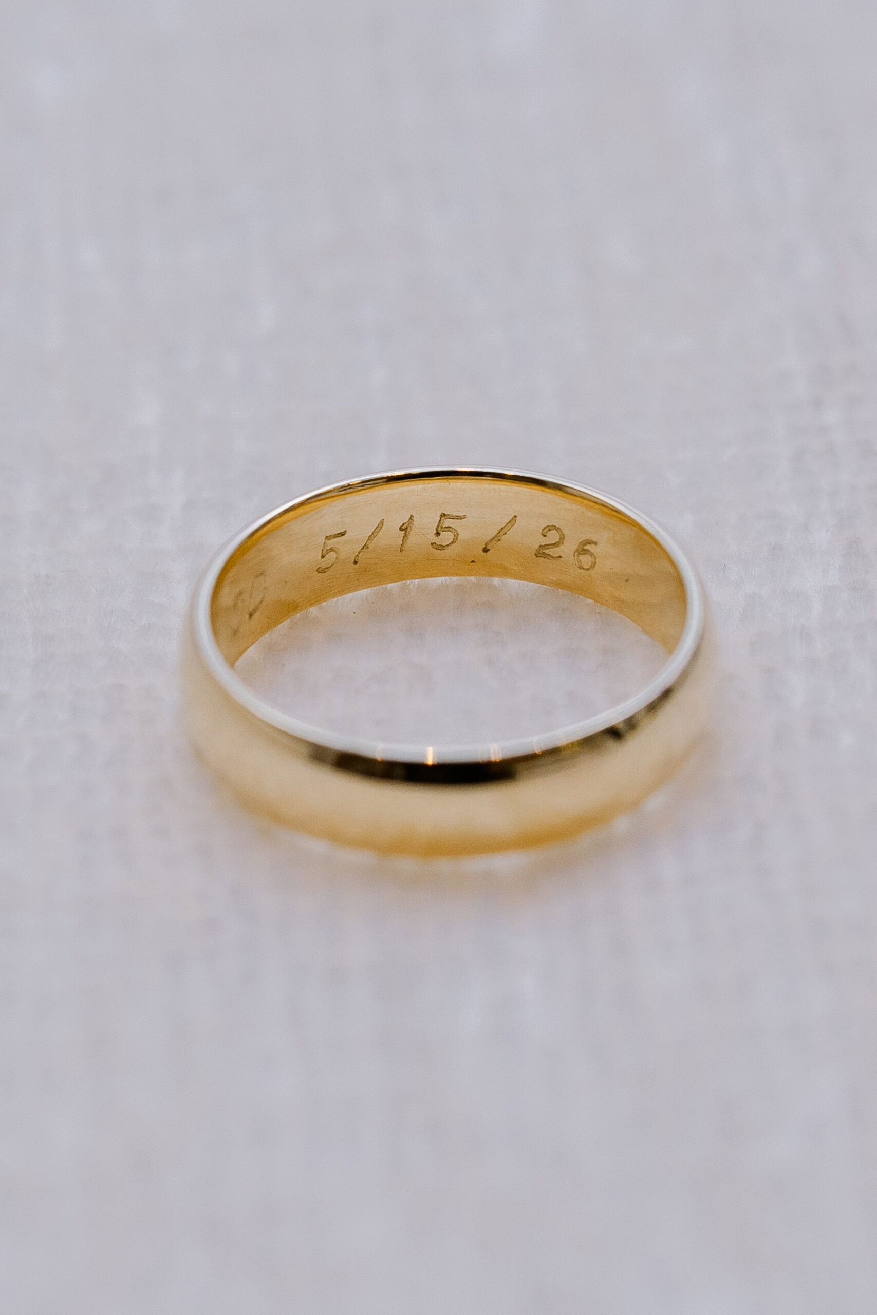 Close-up of a gold wedding ring engraved with the date 5/15/26 on a textured white surface.