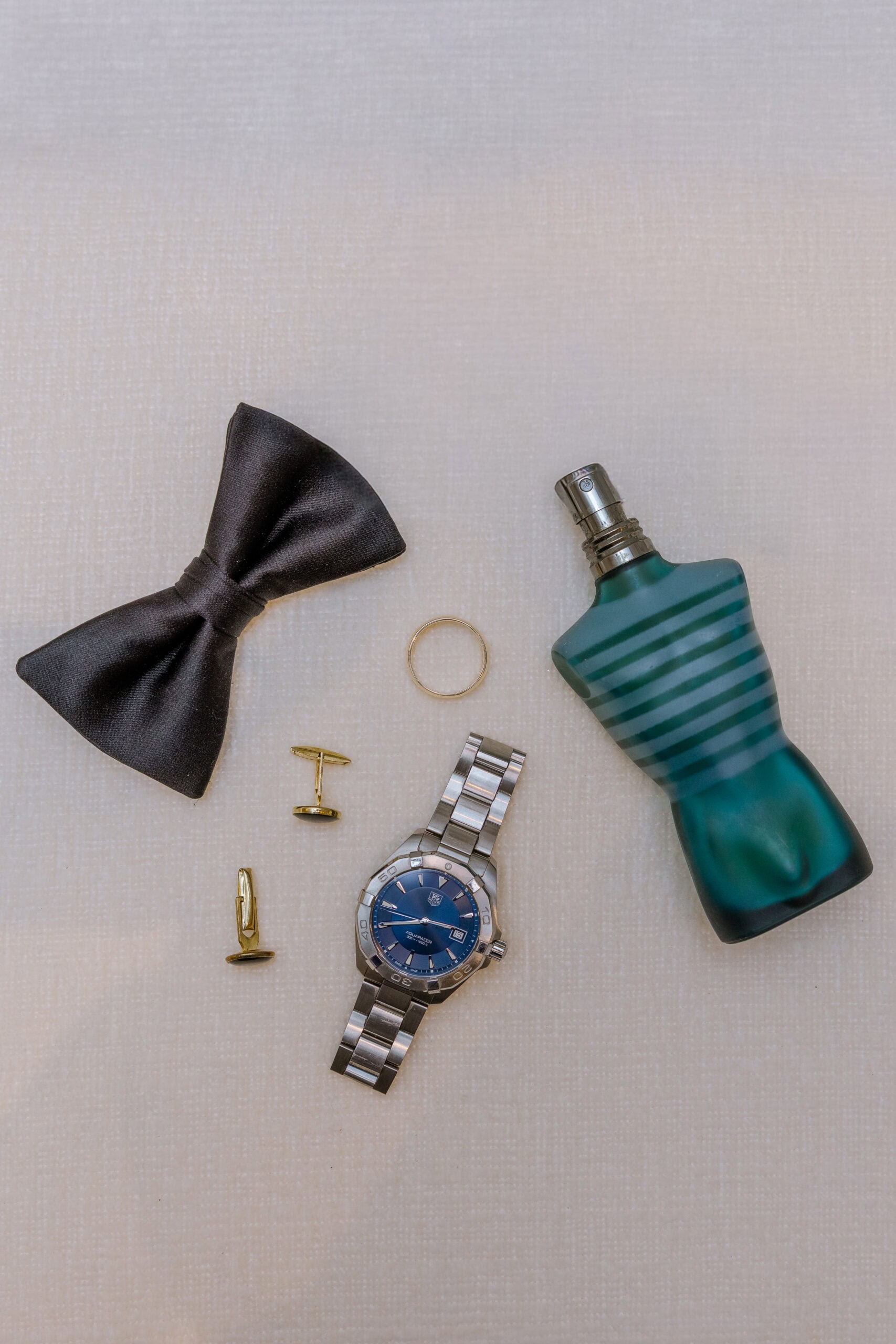 Flat lay of groom's accessories featuring a black bow tie, gold cufflinks, silver wristwatch, wedding band, and green cologne bottle on a beige textured surface.