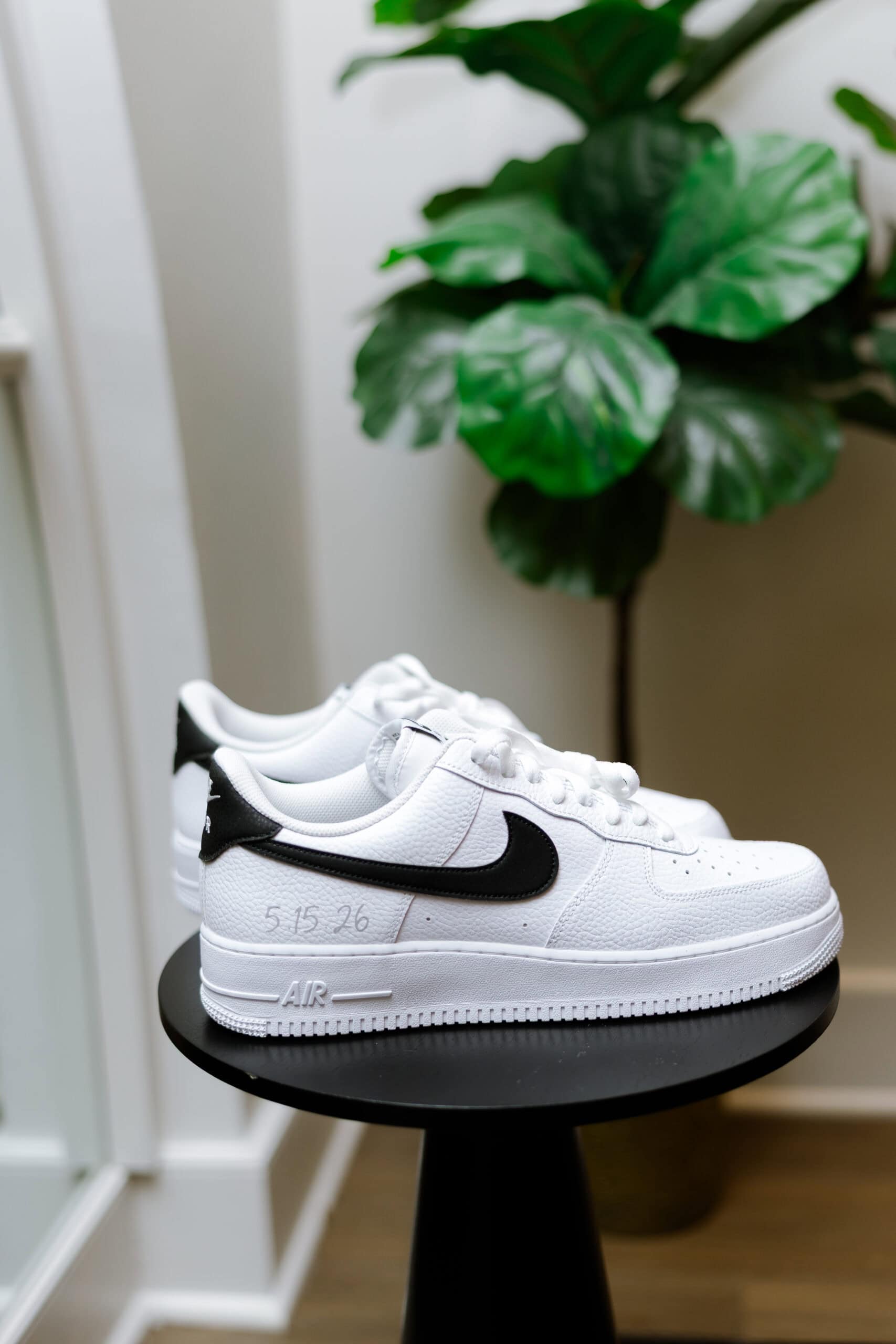 White Nike Air Force 1 sneakers with black swoosh and custom date 5.15.26 on side, placed on black pedestal near green plant.