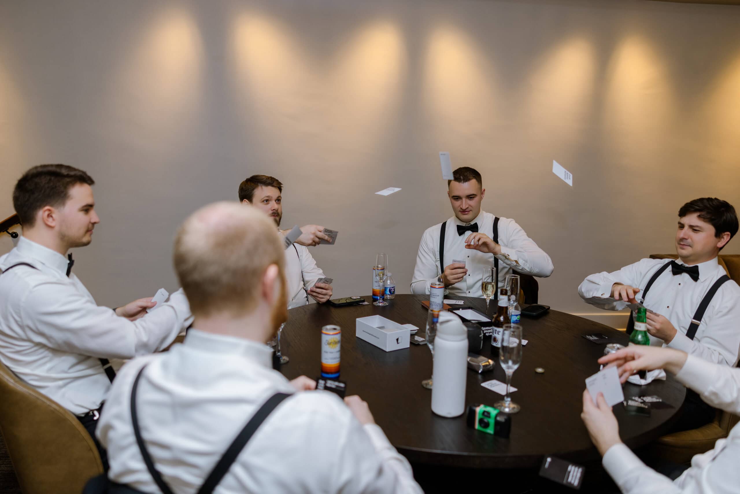 Five men dressed in white shirts with black suspenders and bow ties seated around a table playing a card game, with cards mid-air and drinks on the table, under warm spotlights at Union Trust venue.