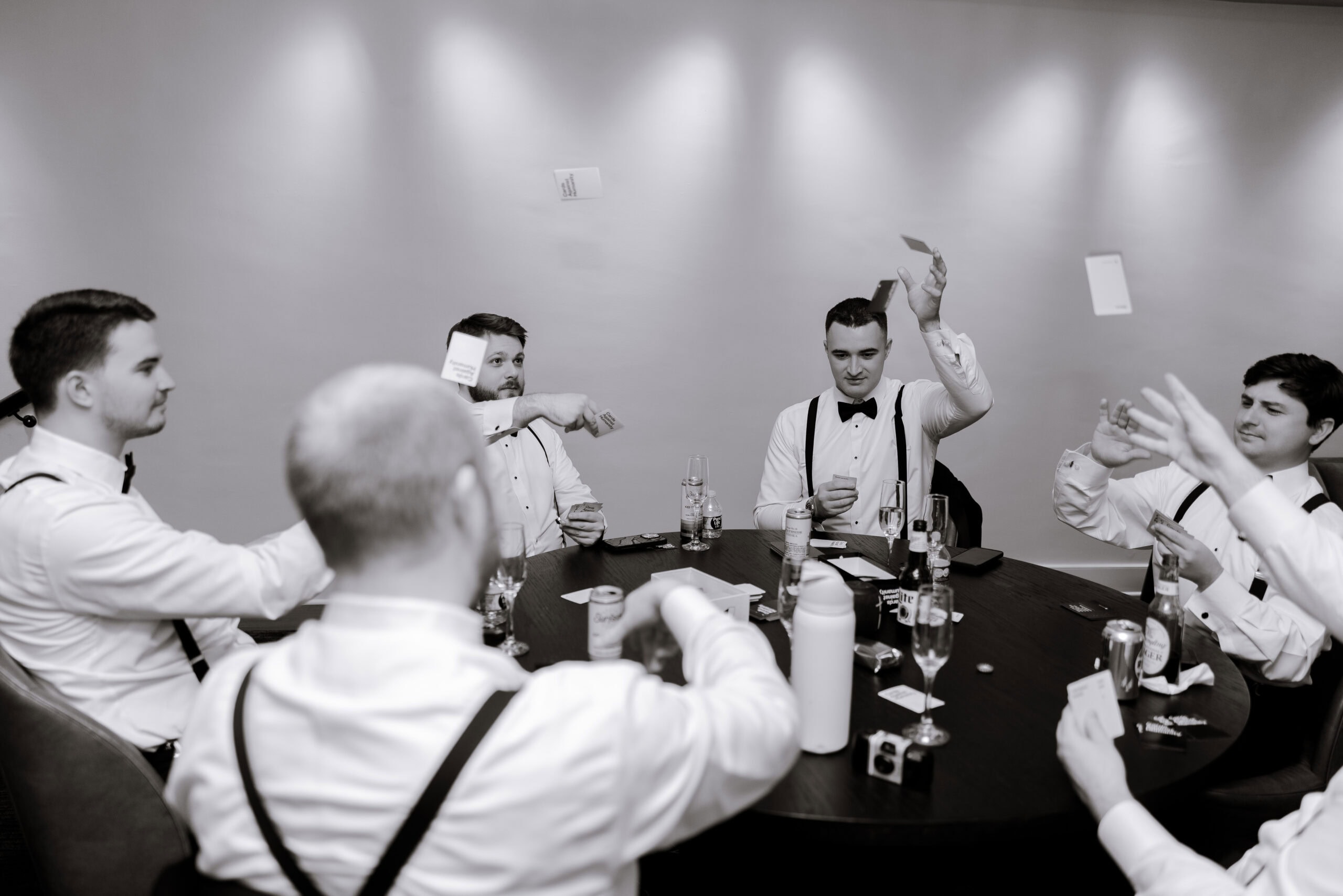 Five men in white shirts and suspenders sit around a round table playing cards, some cards mid-air, in a softly lit room at Union Trust, Philadelphia.