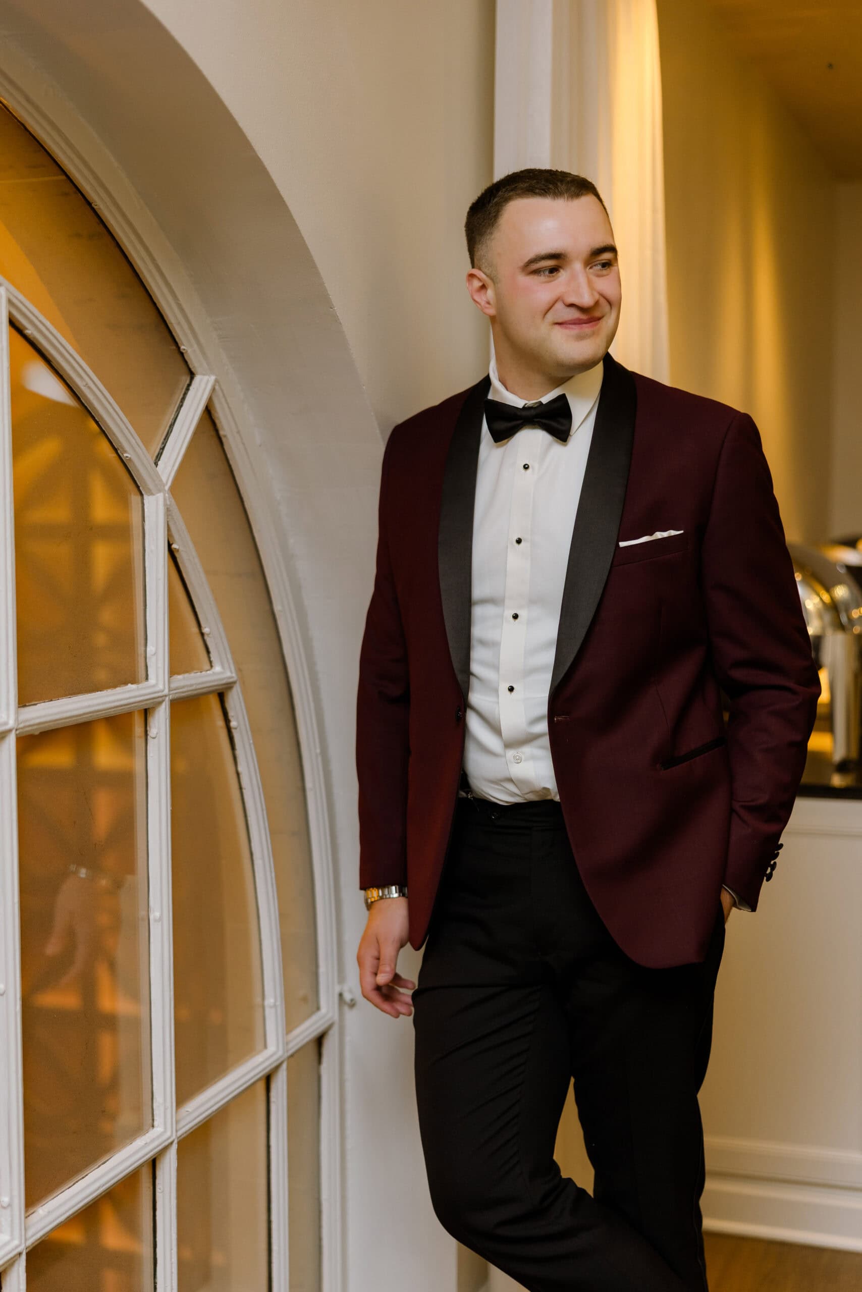 Groom wearing a burgundy tuxedo jacket with black lapels and a black bow tie, standing by an arched window inside Union Trust, Philadelphia.