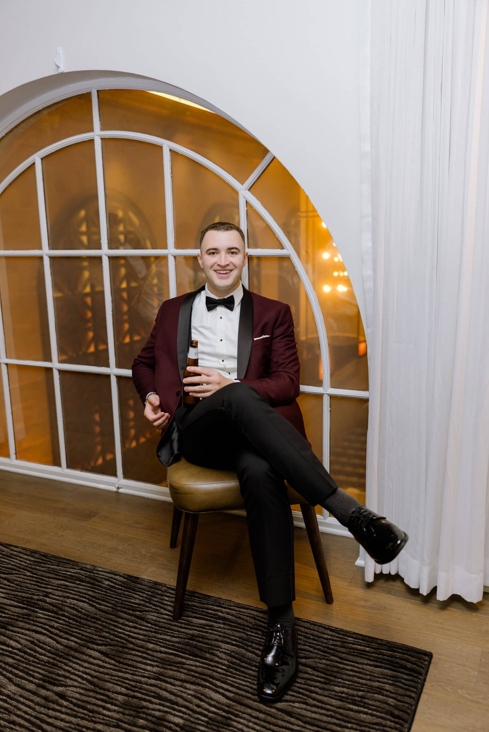 Man in burgundy tuxedo jacket and black bow tie sitting on a stool by an arched window holding a bottle, indoors at Union Trust Philadelphia.