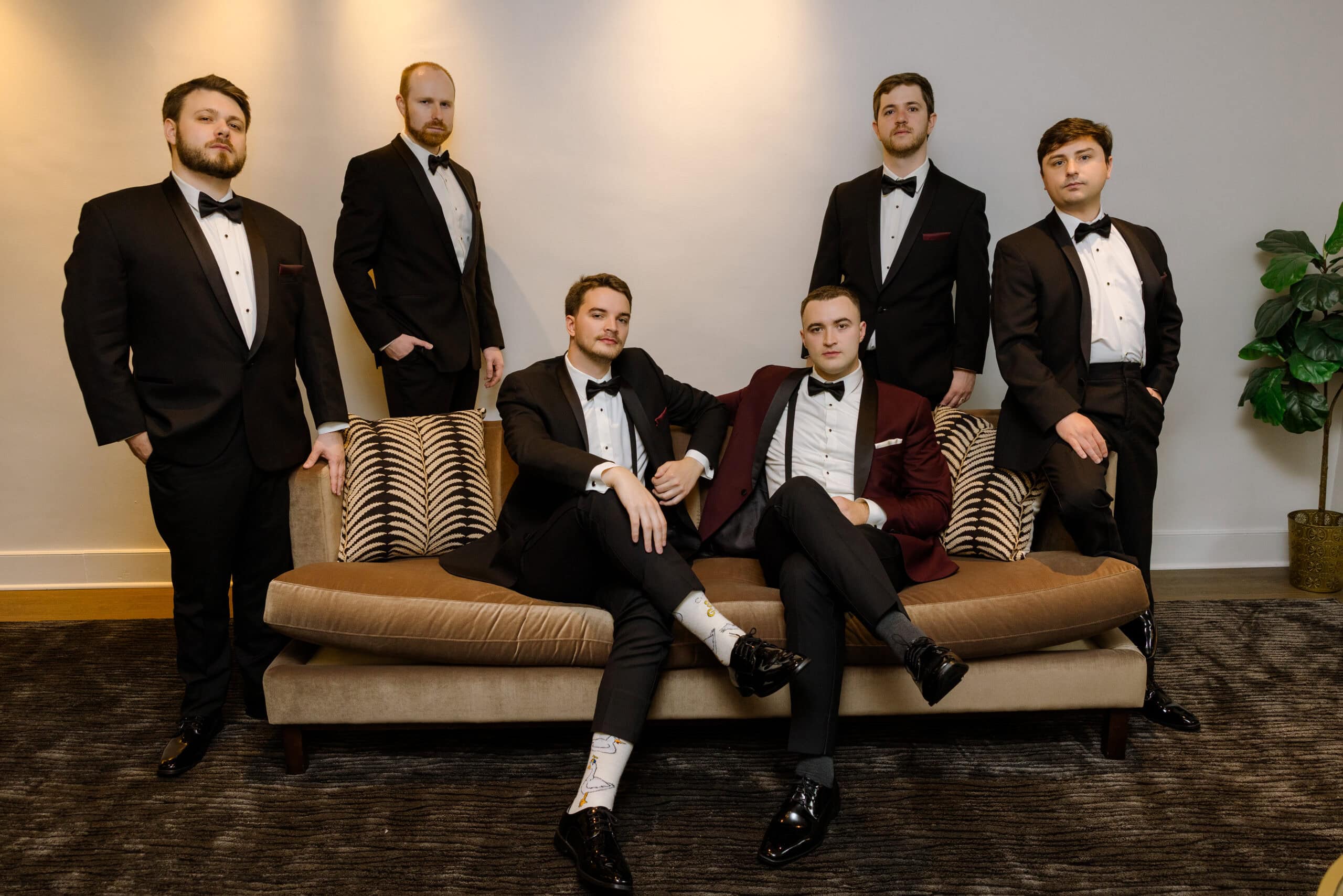 Six men in tuxedos posing indoors on and around a beige sofa, against a neutral wall with soft lighting, at Union Trust venue.