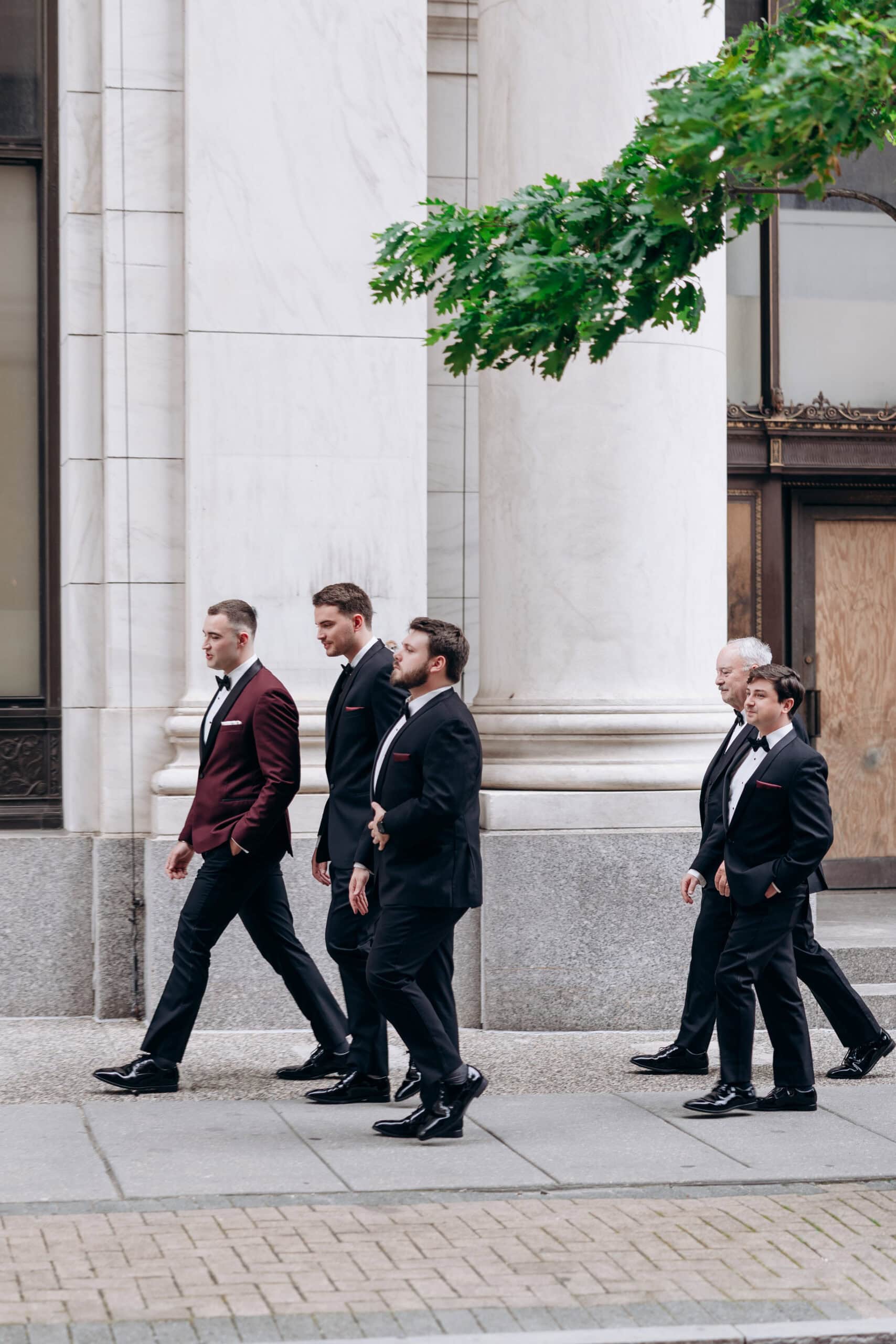 Five groomsmen in tuxedos walking in a line beside large white marble columns with green leaves overhead at Union Trust, Philadelphia.