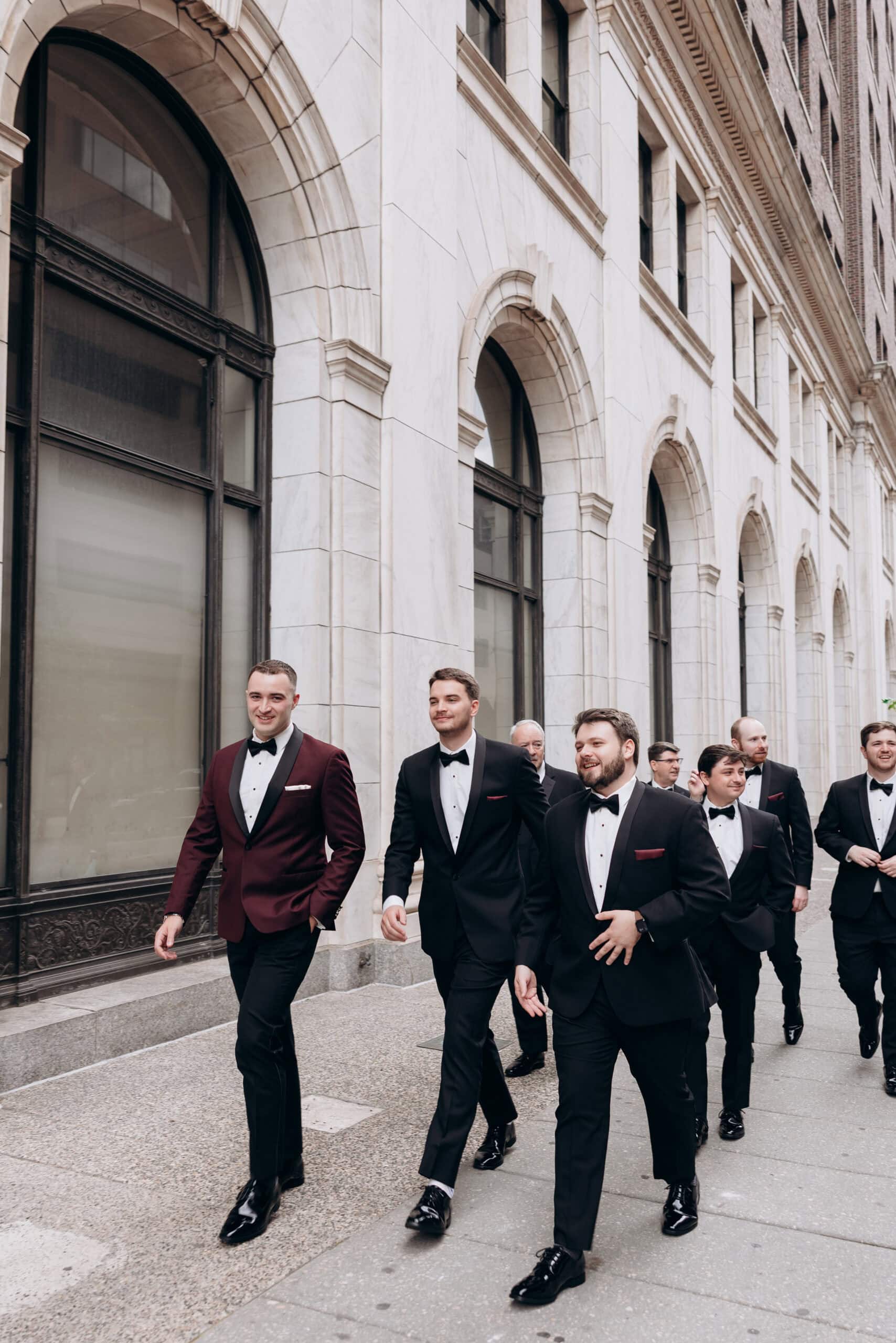 Group of groomsmen in black and burgundy tuxedos walking on sidewalk beside marble building with arched windows at Union Trust, Philadelphia.