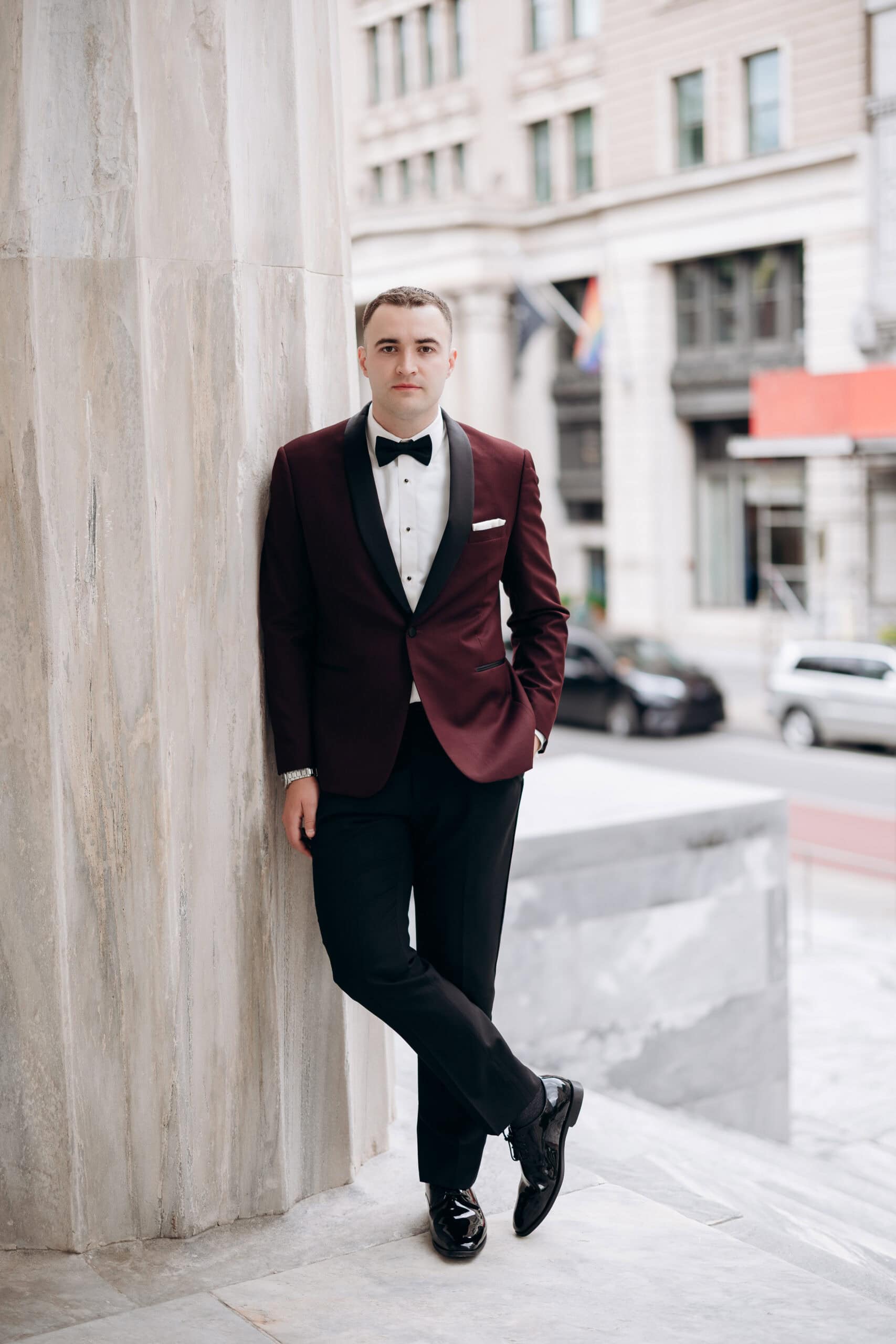 Man in burgundy tuxedo with black lapels leaning against marble column outside Union Trust building in Philadelphia.