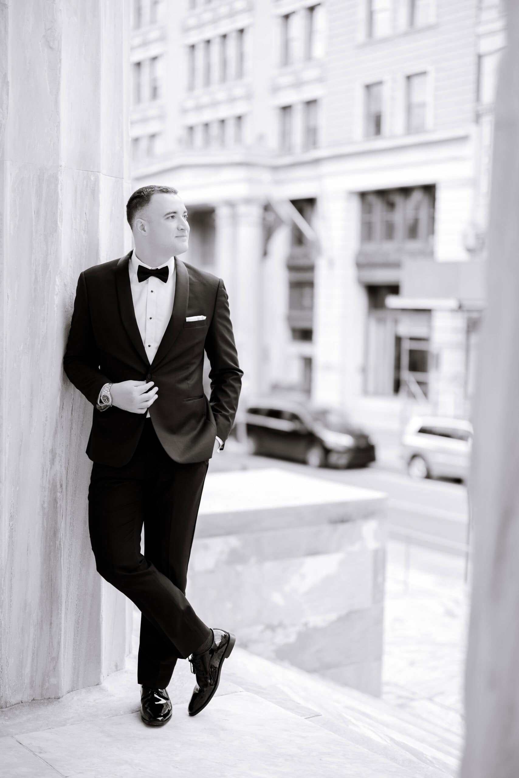 Man in black tuxedo with bow tie leaning against marble column, urban building facade blurred in background, daytime