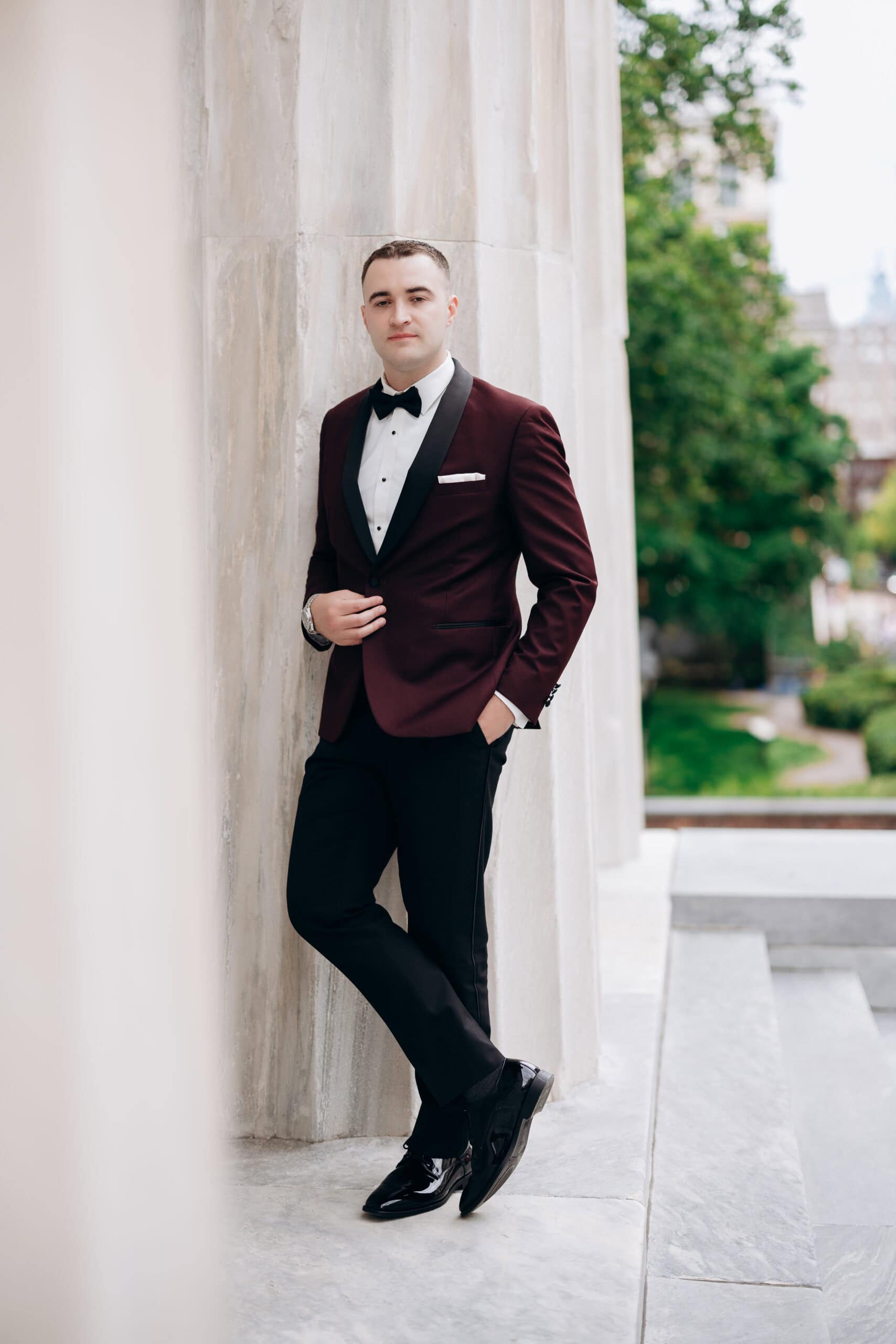 Man in burgundy tuxedo with black lapels and bow tie leaning against a marble column outside Union Trust, Philadelphia