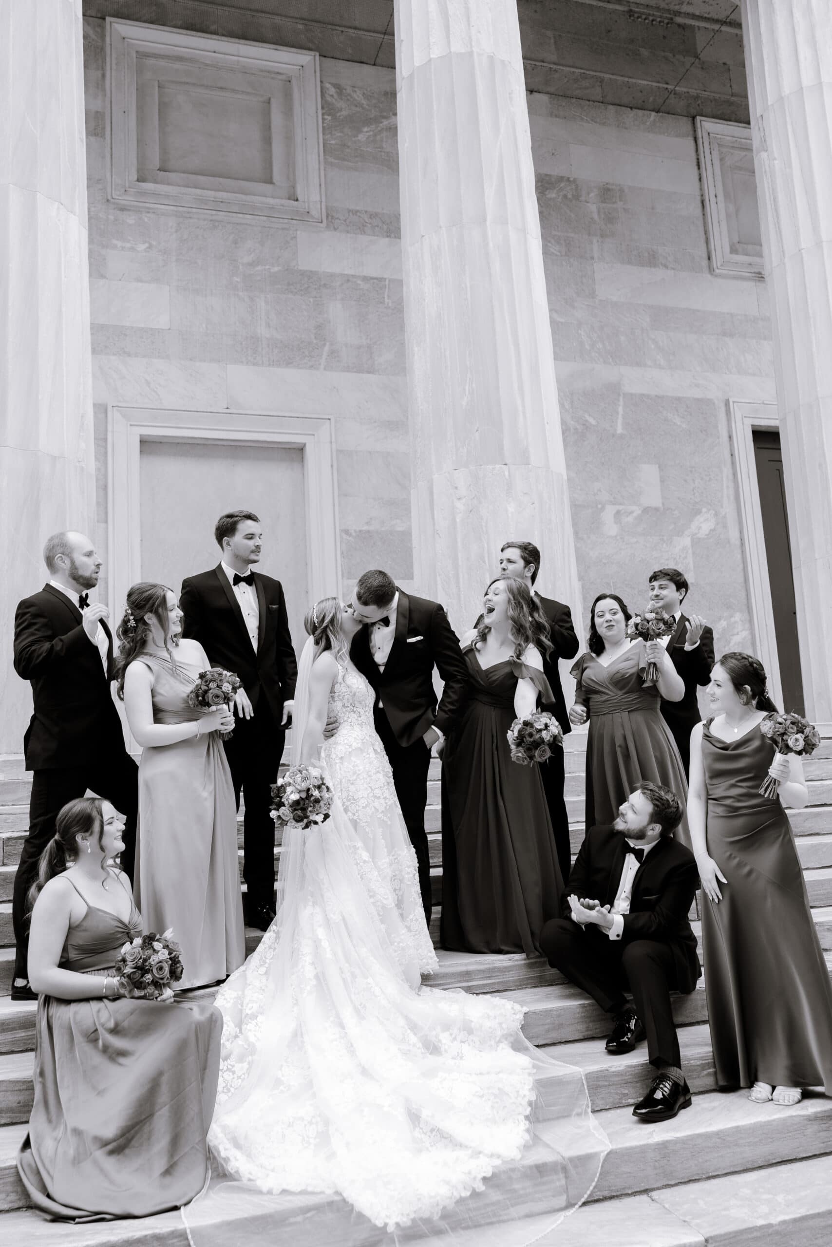 Wedding party in formal attire with bride and groom kissing on stone steps framed by large marble columns at Union Trust, Philadelphia.