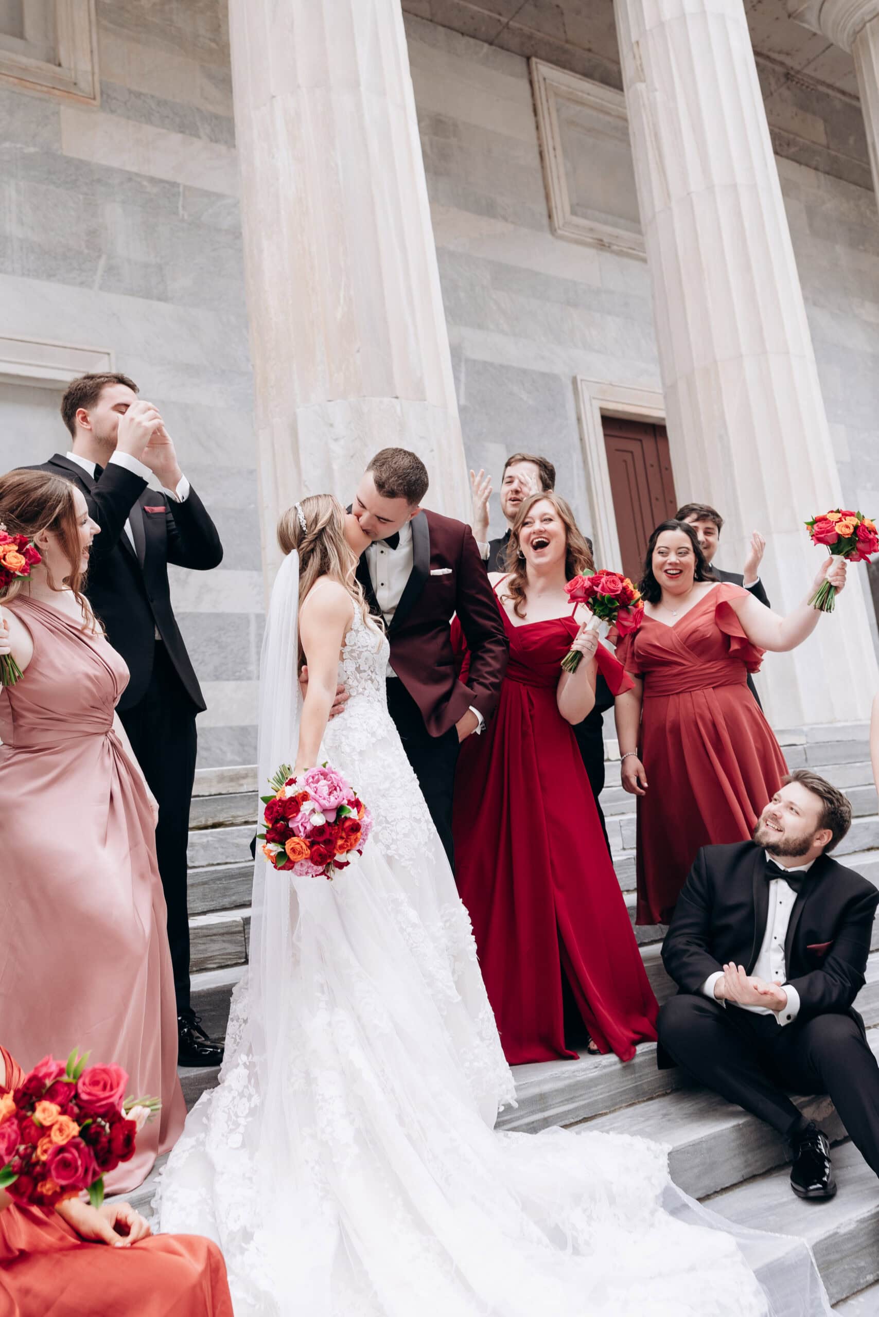 Bride and groom kissing on marble steps with bridal party in red and blush dresses, celebrating outside Union Trust, Philadelphia.
