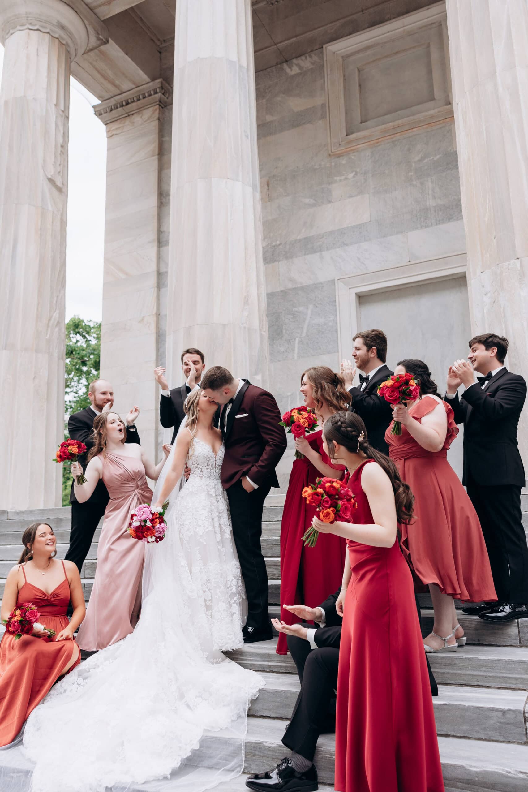 Bride and groom kissing on marble steps with bridesmaids in red and blush dresses and groomsmen in black tuxedos, Union Trust Philadelphia.