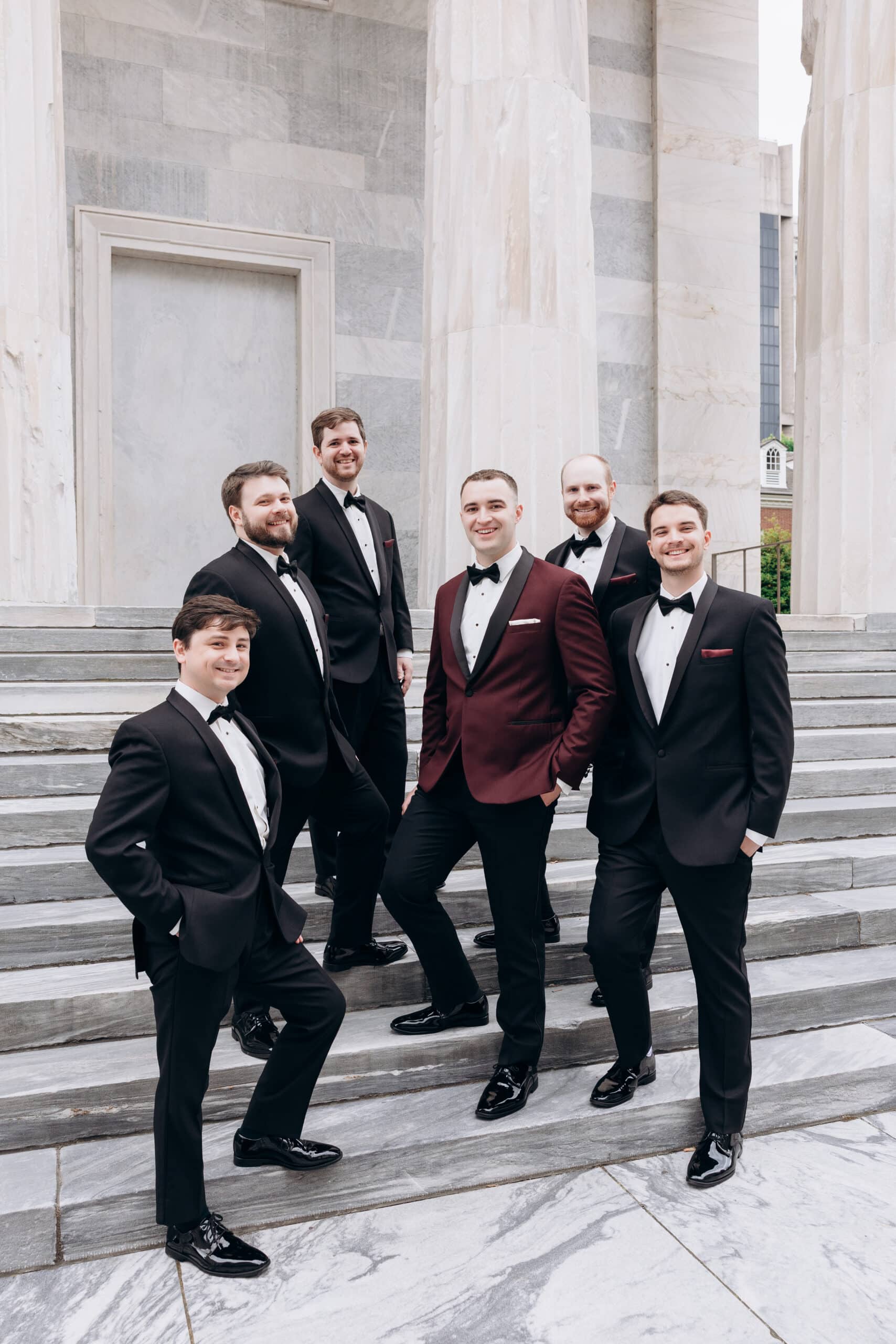 Six groomsmen wearing black tuxedos and one in a burgundy tuxedo jacket standing on marble steps with tall marble columns in the background at Union Trust, Philadelphia.