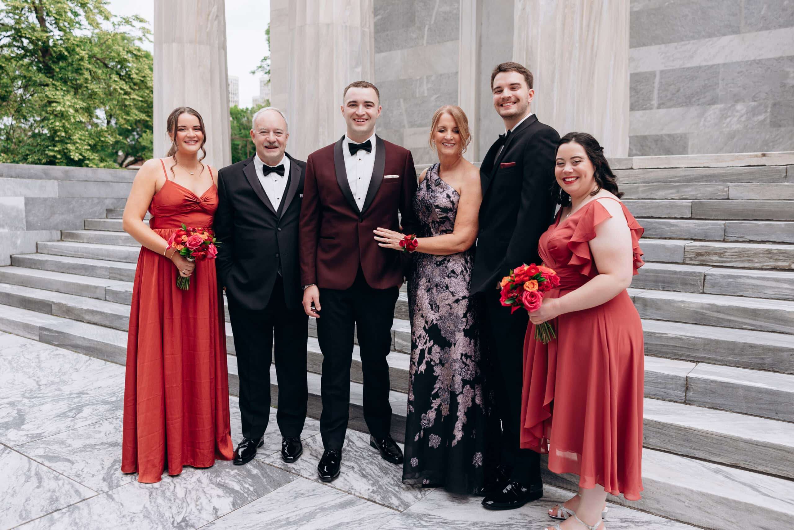Wedding party in formal attire standing on marble steps with columns at Union Trust Philadelphia, featuring red gowns and tuxedos.