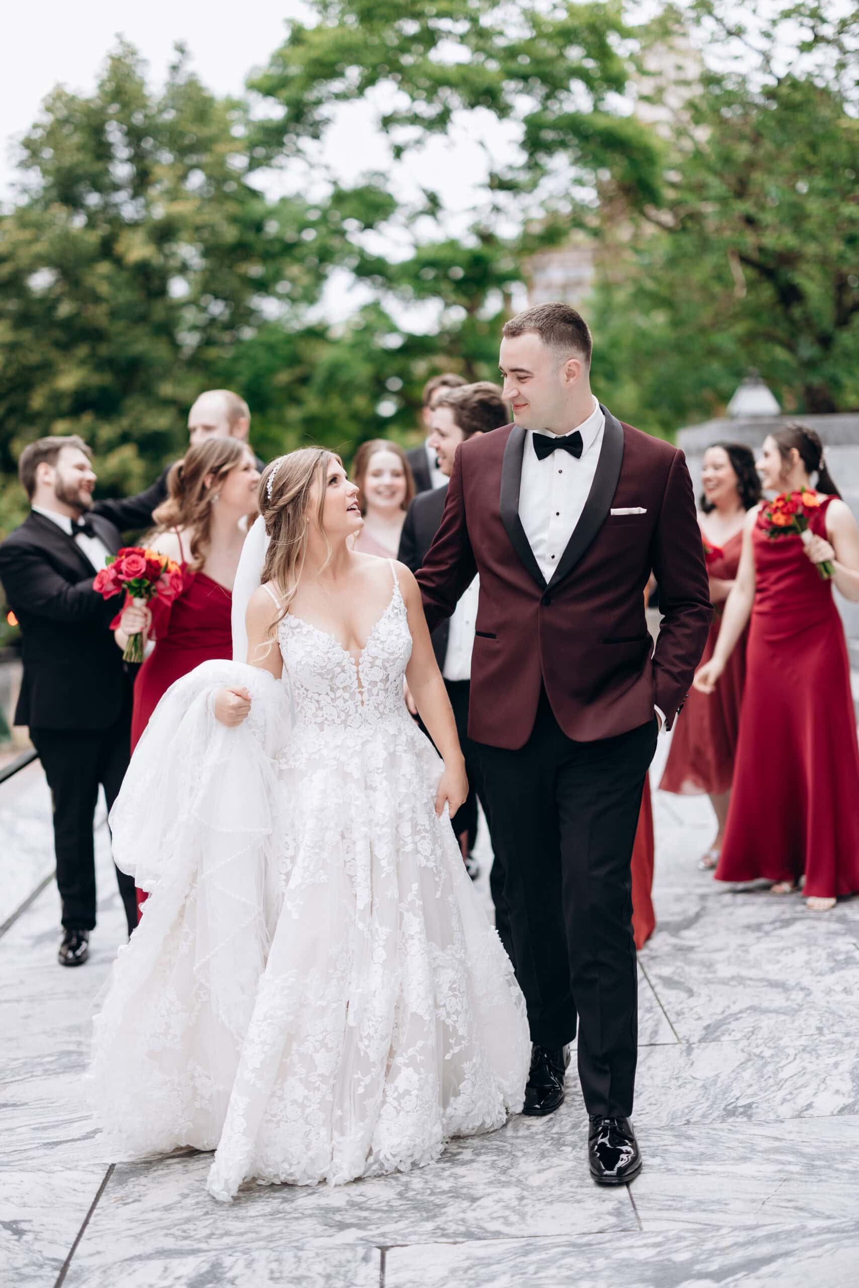 Bride in detailed lace wedding dress and groom in burgundy tuxedo walking and looking at each other, bridal party in red dresses and black tuxedos behind them, outdoors with greenery.