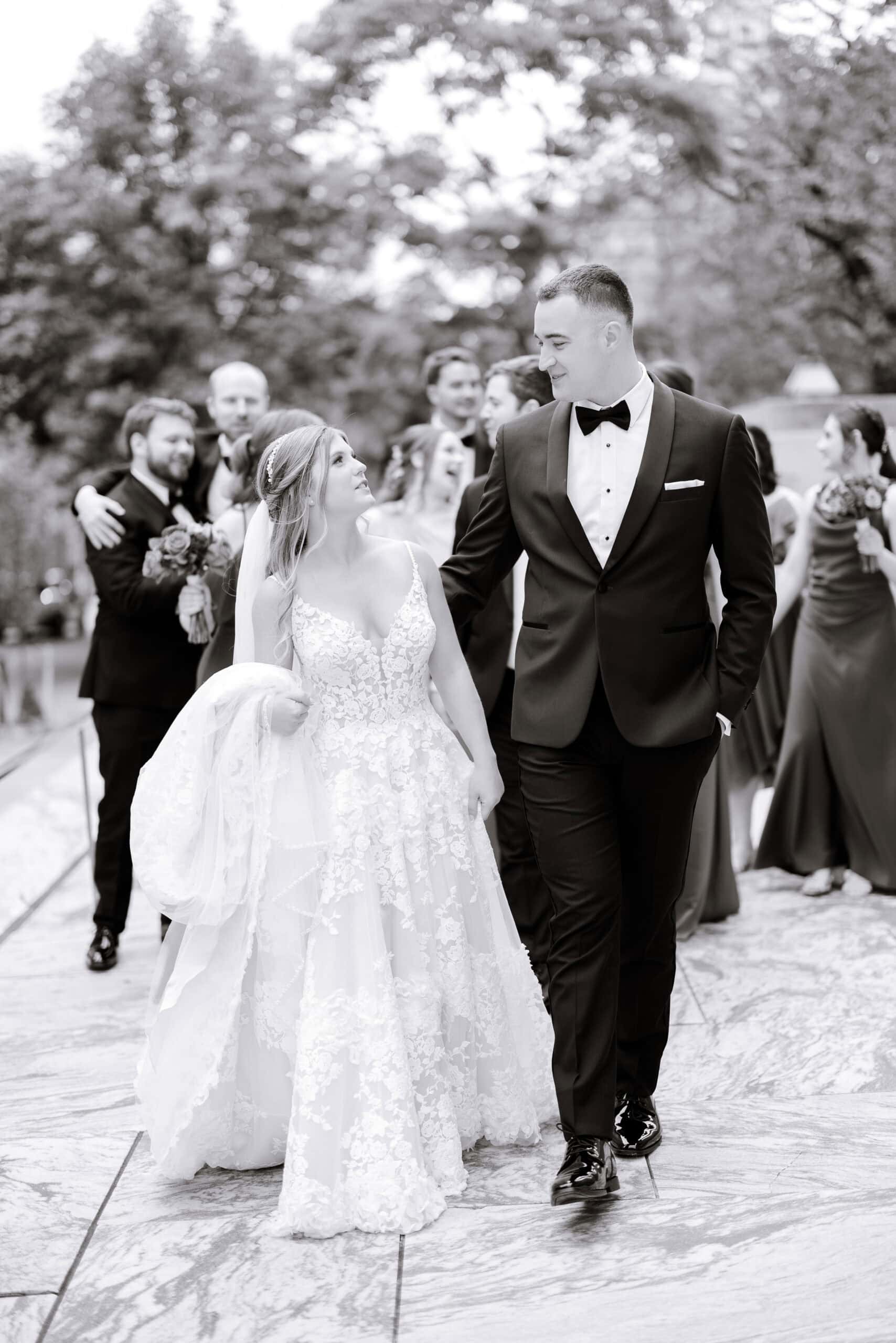 Bride in lace wedding dress and groom in tuxedo walking hand in hand with bridal party in the background, outdoors on marble pavement.
