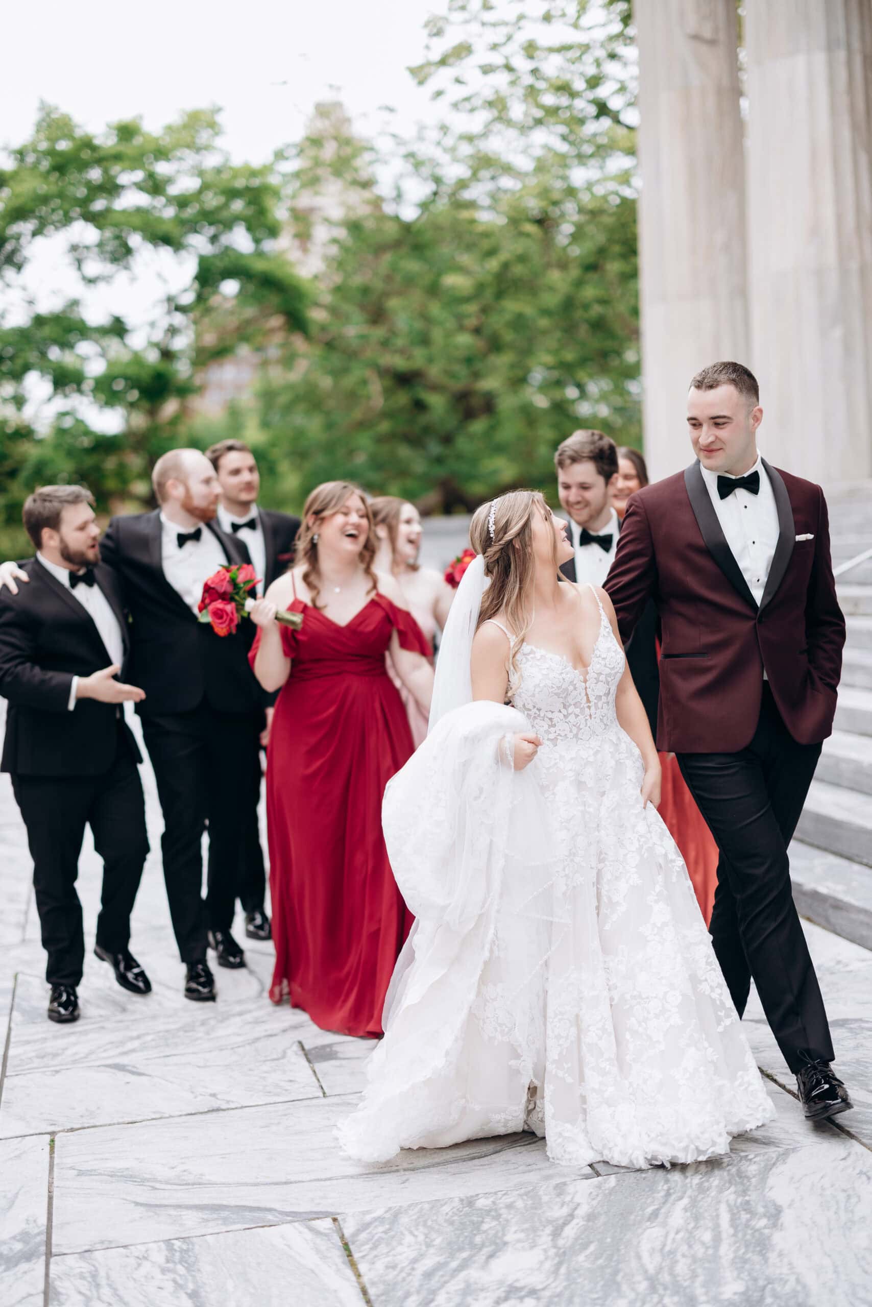 Bride in lace wedding dress and groom in burgundy tuxedo walk with bridal party in black tuxedos and red dresses outside Union Trust, Philadelphia.