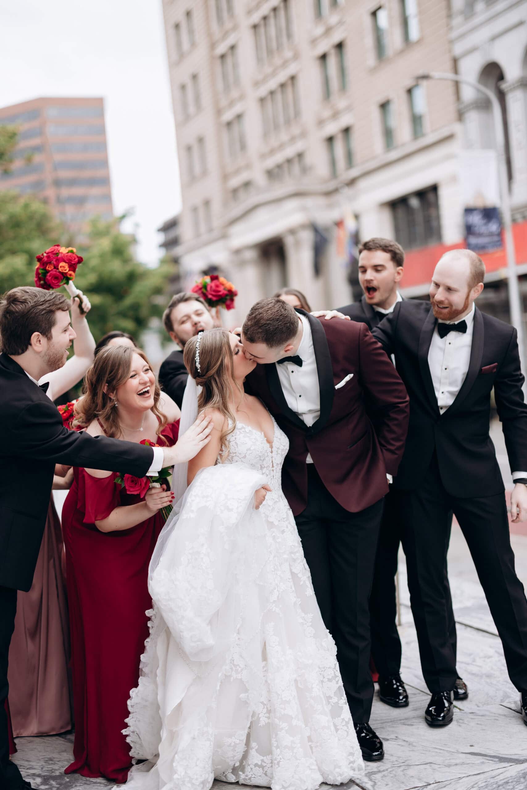 Bride in lace wedding dress and groom in burgundy tuxedo kiss while surrounded by joyful bridal party in red and black formal wear on city sidewalk.