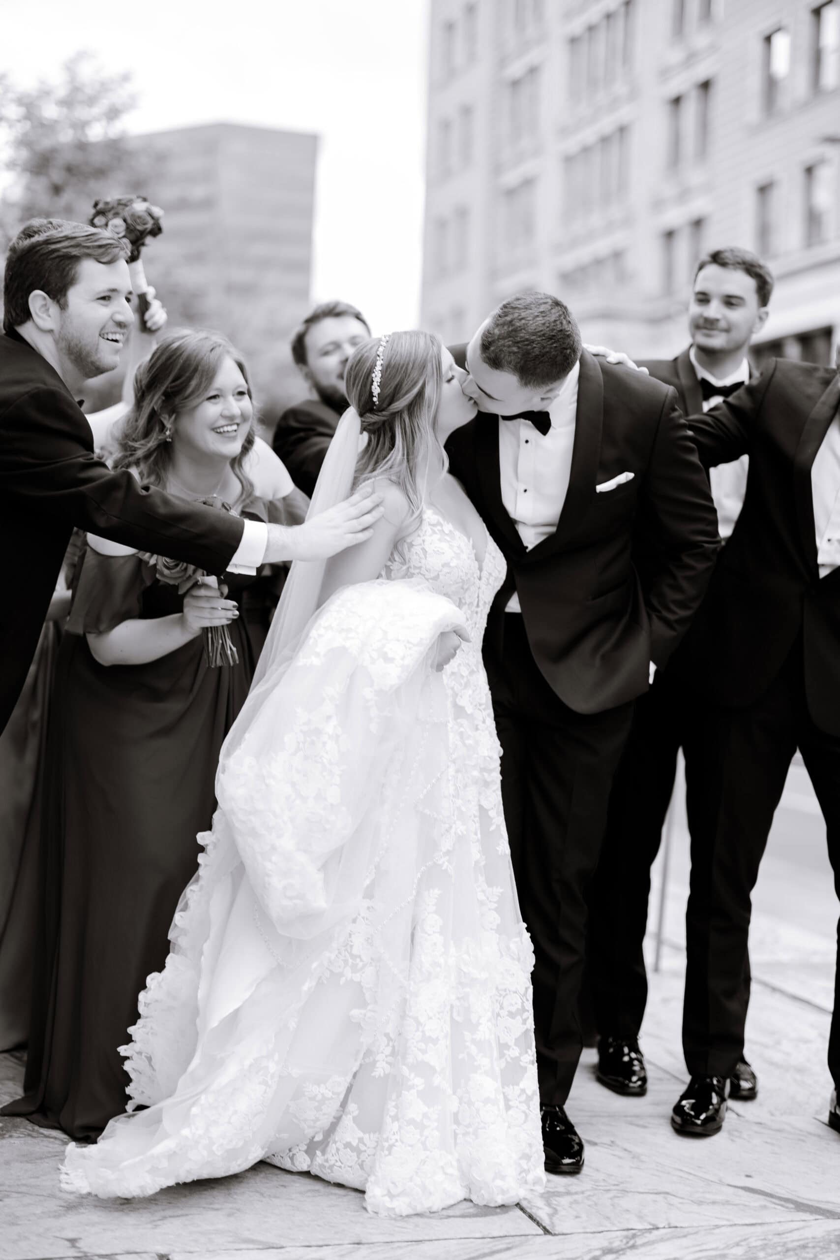 Black and white photo of a bride and groom kissing surrounded by smiling bridal party members in formal attire on a city street at Union Trust, Philadelphia.