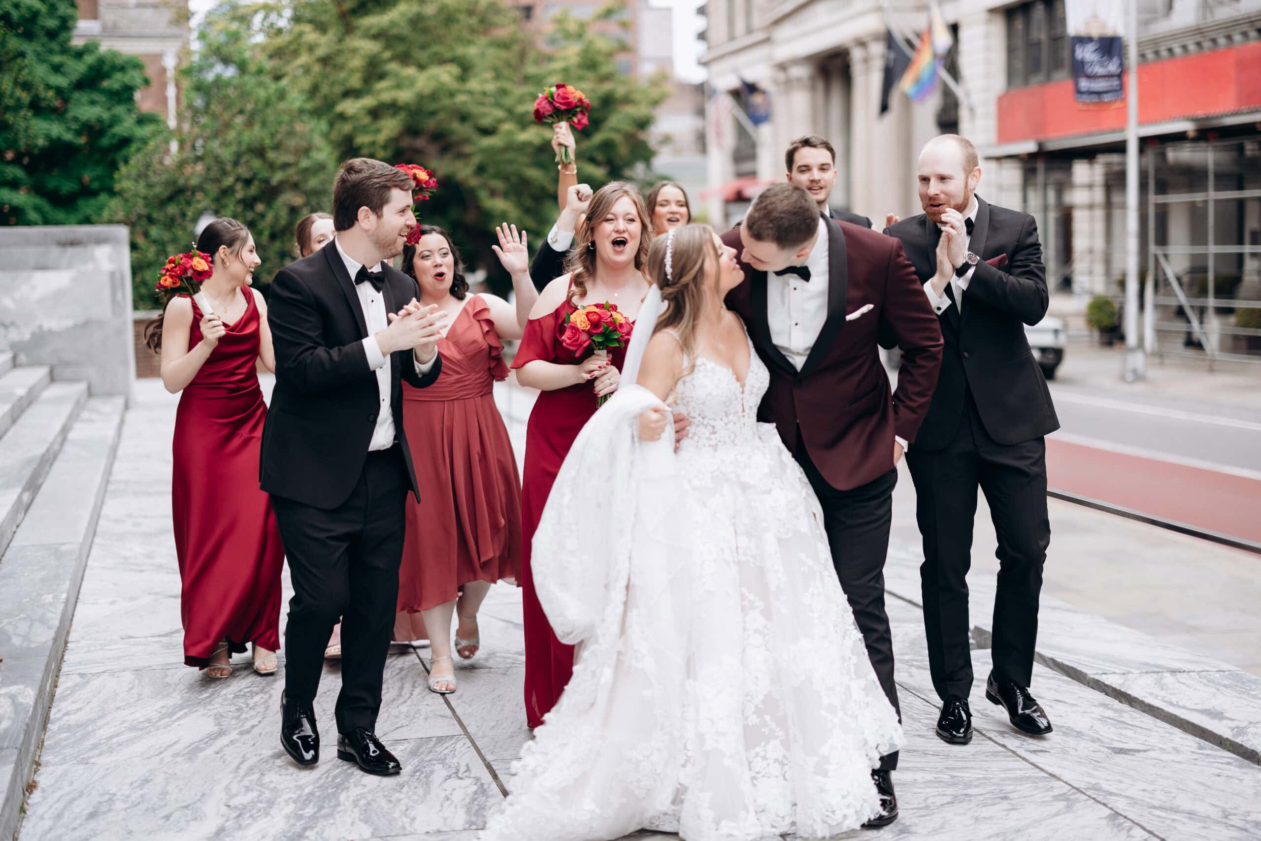 Wedding party in burgundy dresses and black tuxedos celebrating as groom leans in to kiss bride in white lace gown on city sidewalk.