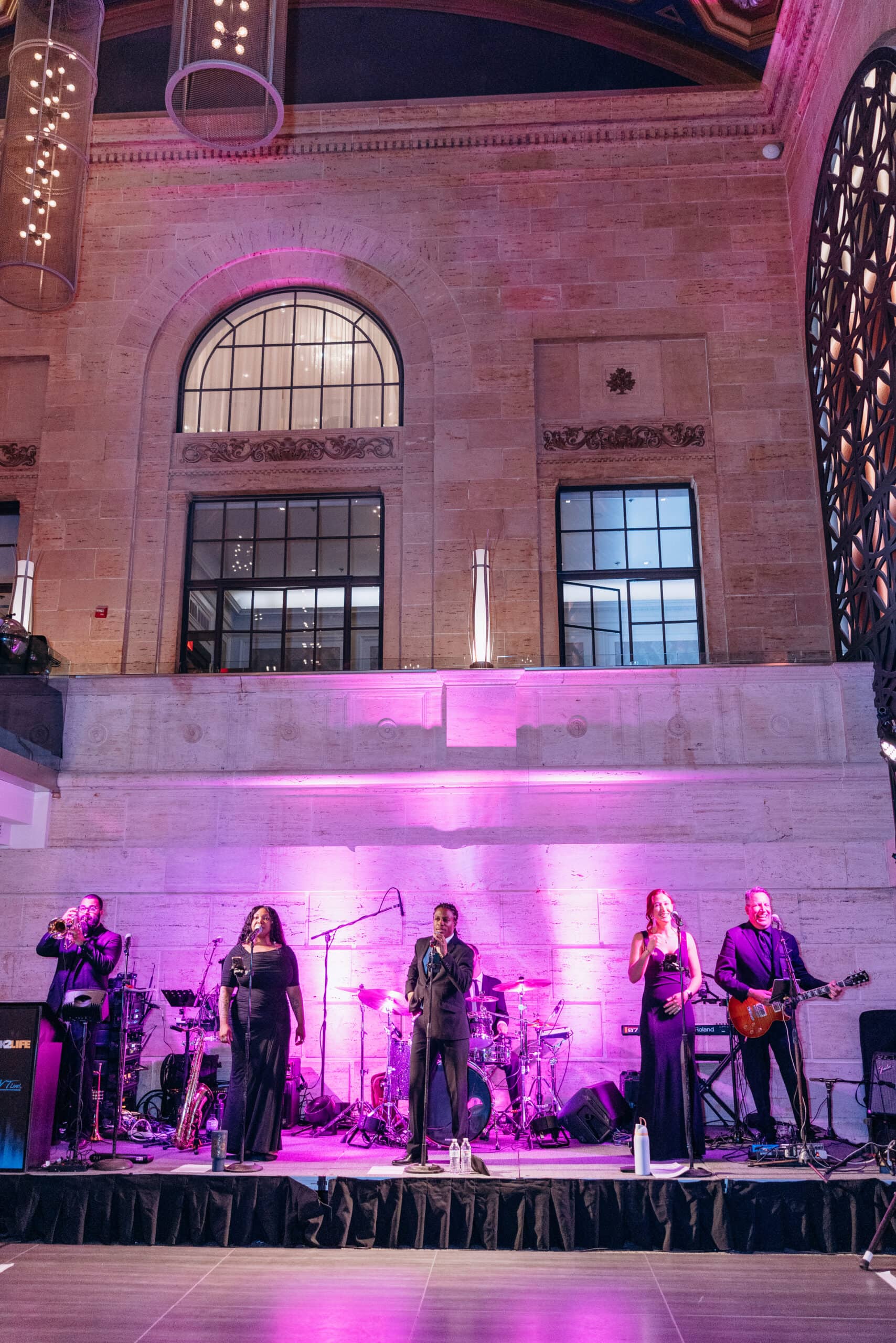 Five-piece jazz band performing on stage with trumpet, vocals, guitar, and drums in front of large arched windows and stone walls illuminated with purple light at Union Trust, Philadelphia.