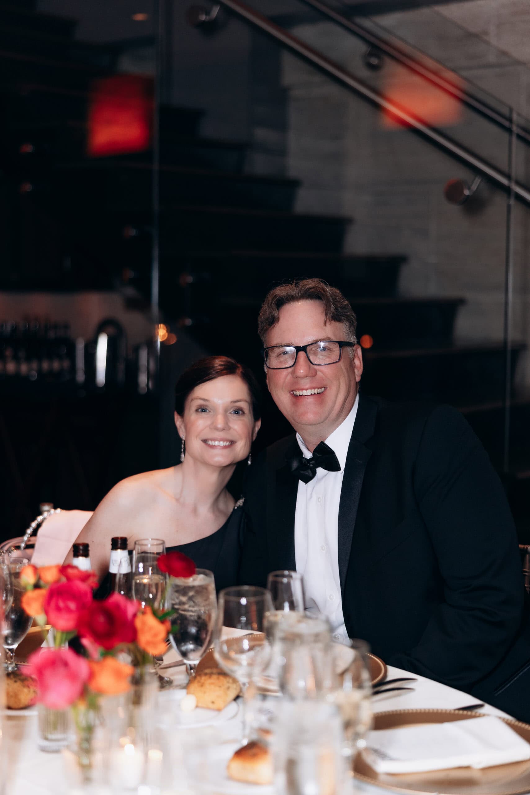 Man and woman dressed formally, seated at a wedding reception table with glasses, plates, and red-orange floral centerpiece, dark staircase in background at Union Trust, Philadelphia.