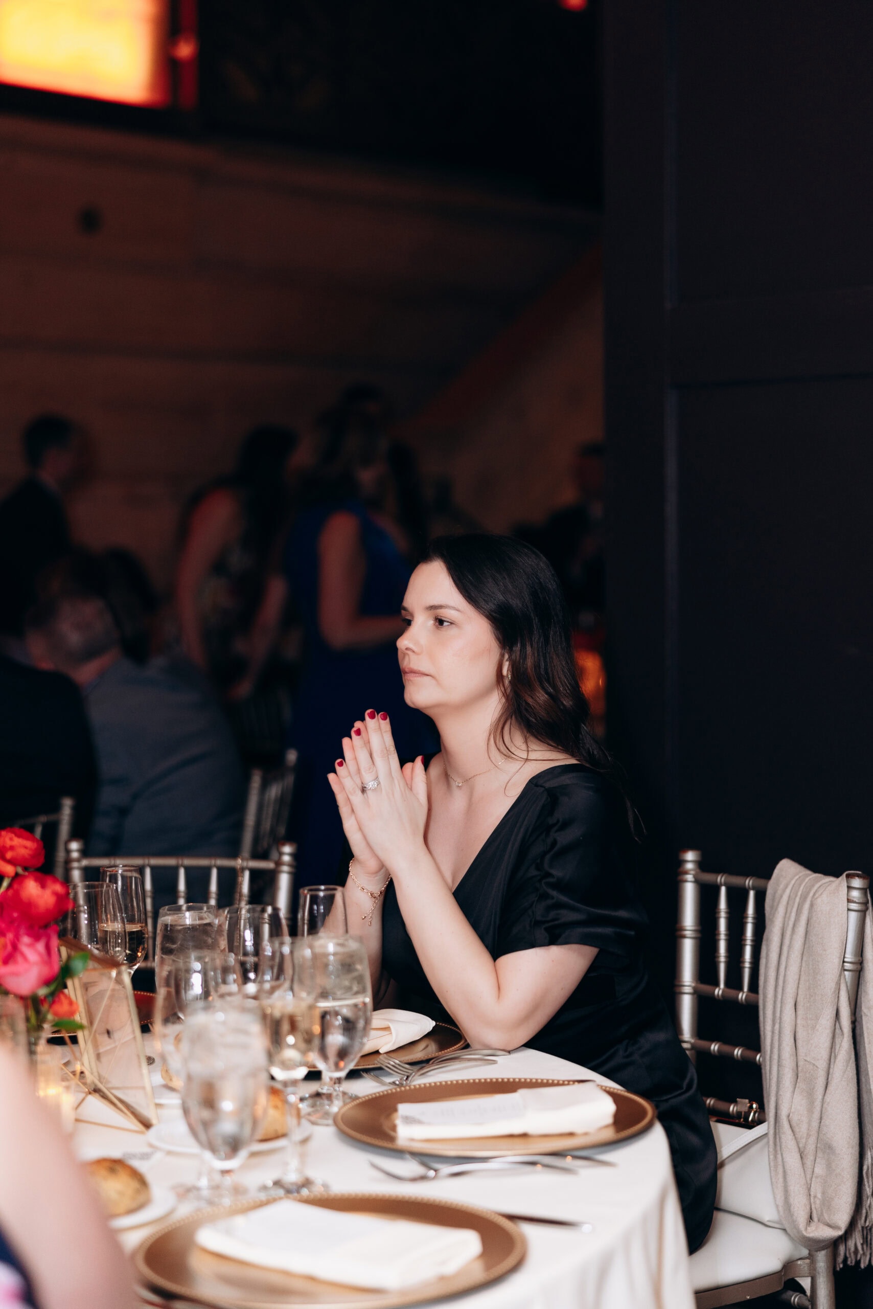 Woman in black dress with hands clasped, seated at a formal wedding table with gold-rimmed plates and glassware, Union Trust venue.
