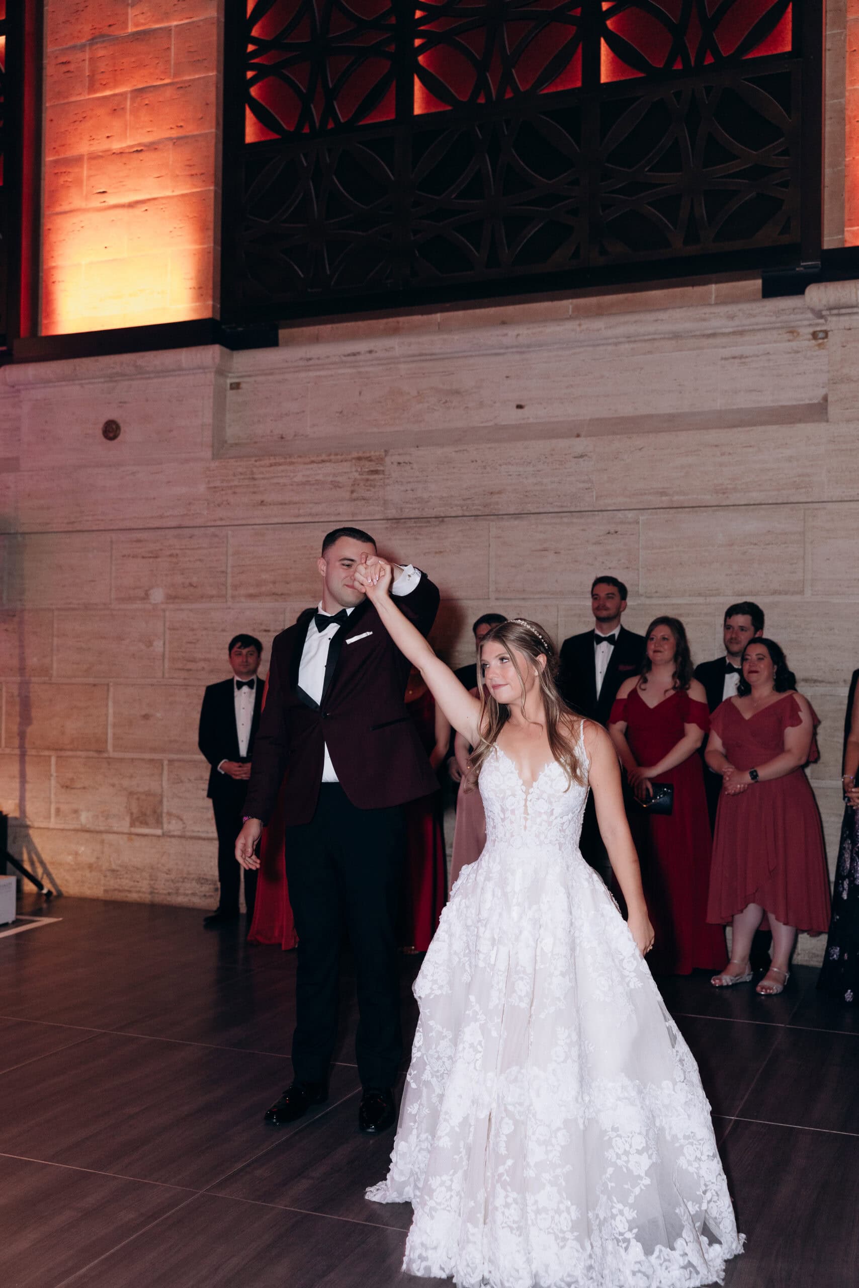 Bride and groom sharing an intimate first dance at Union Trust Philadelphia with guests watching, groom in burgundy tuxedo, bride in floral lace gown.
