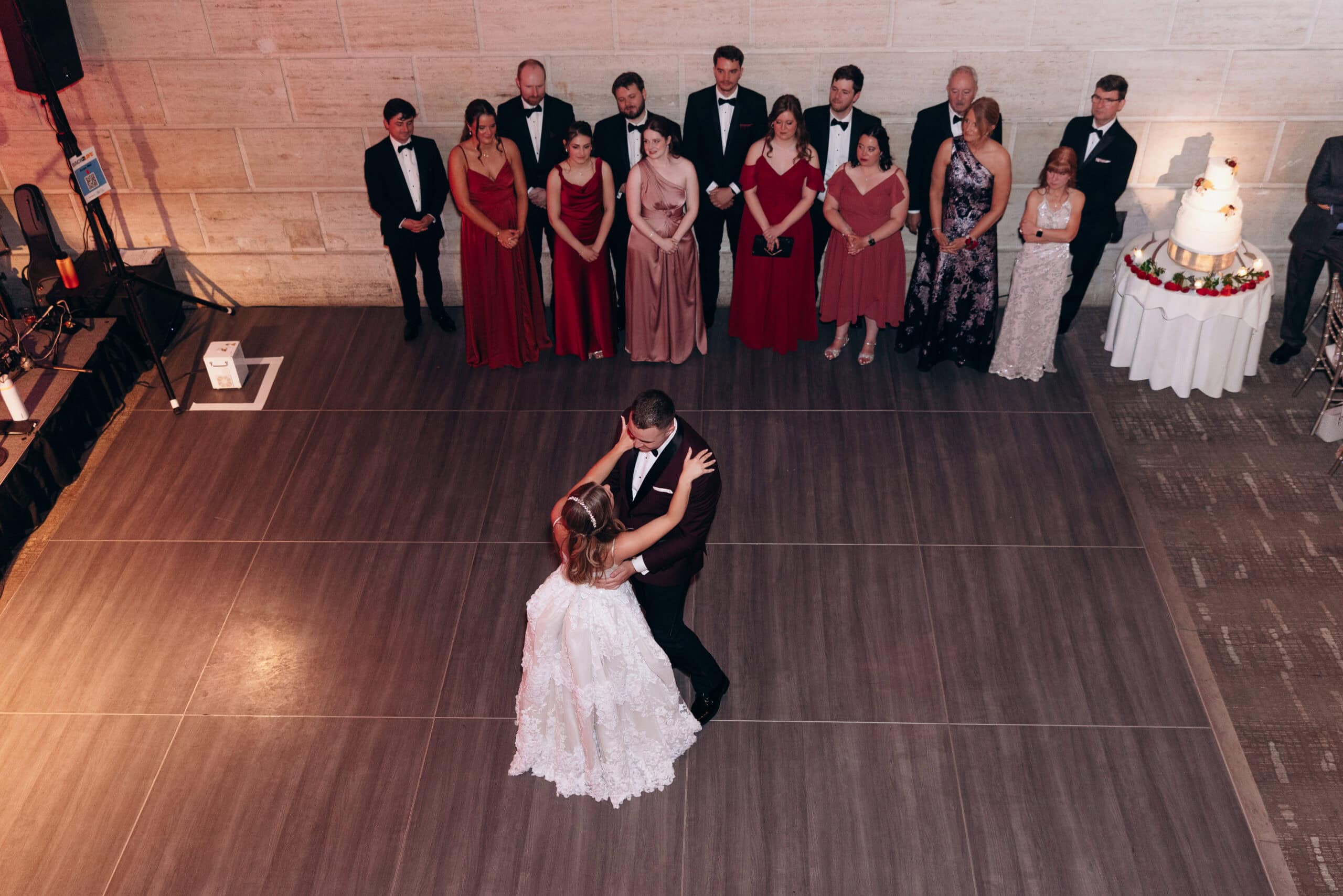 Overhead view of bride and groom sharing first dance on wooden floor, surrounded by bridesmaids in red and blush gowns and groomsmen in tuxedos, with a white tiered wedding cake on a round table nearby at Union Trust, Philadelphia.
