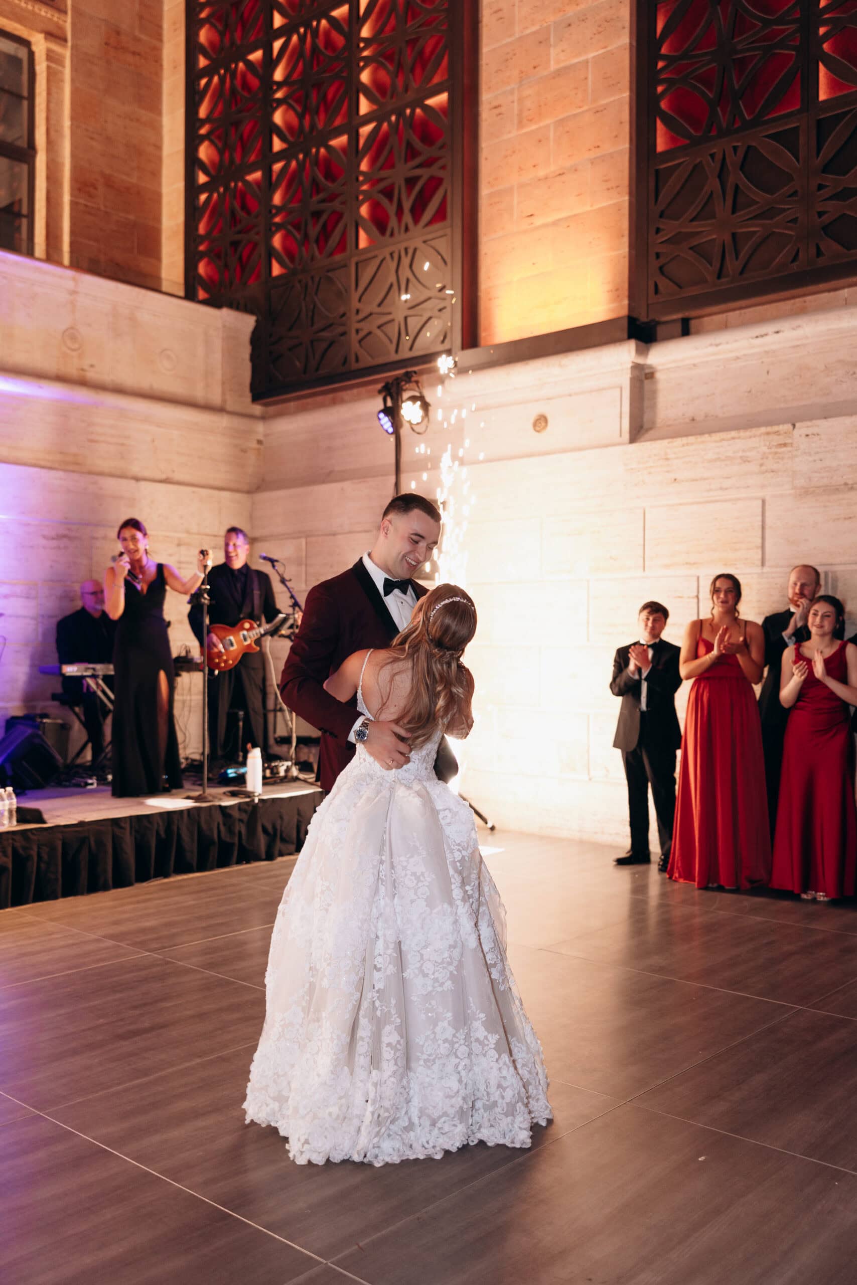Bride and groom share their first dance in an elegant hall with stone walls and decorative iron panels, surrounded by musicians and applauding guests.