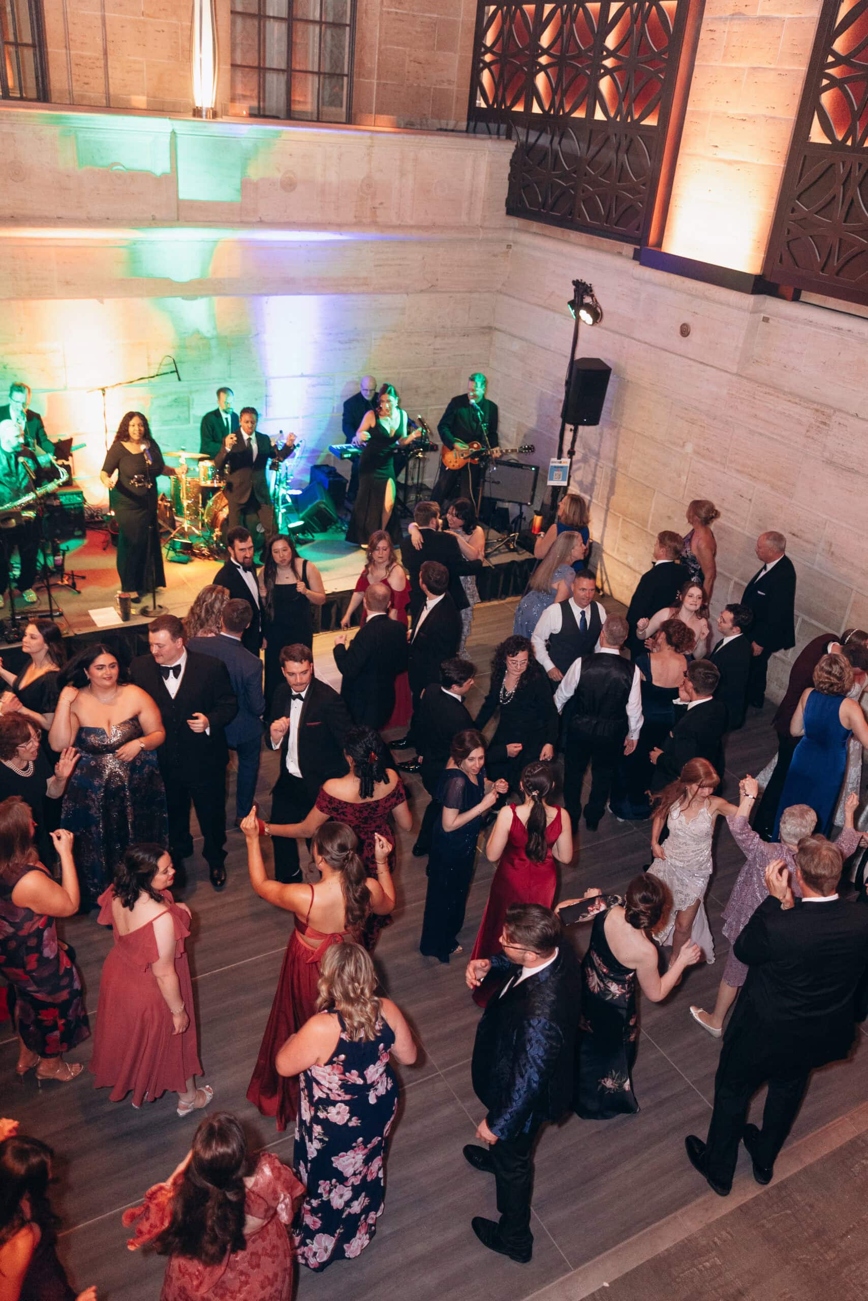 Overhead view of guests in formal attire dancing to a live band performing on a small stage in a stone-walled venue with colorful uplighting.