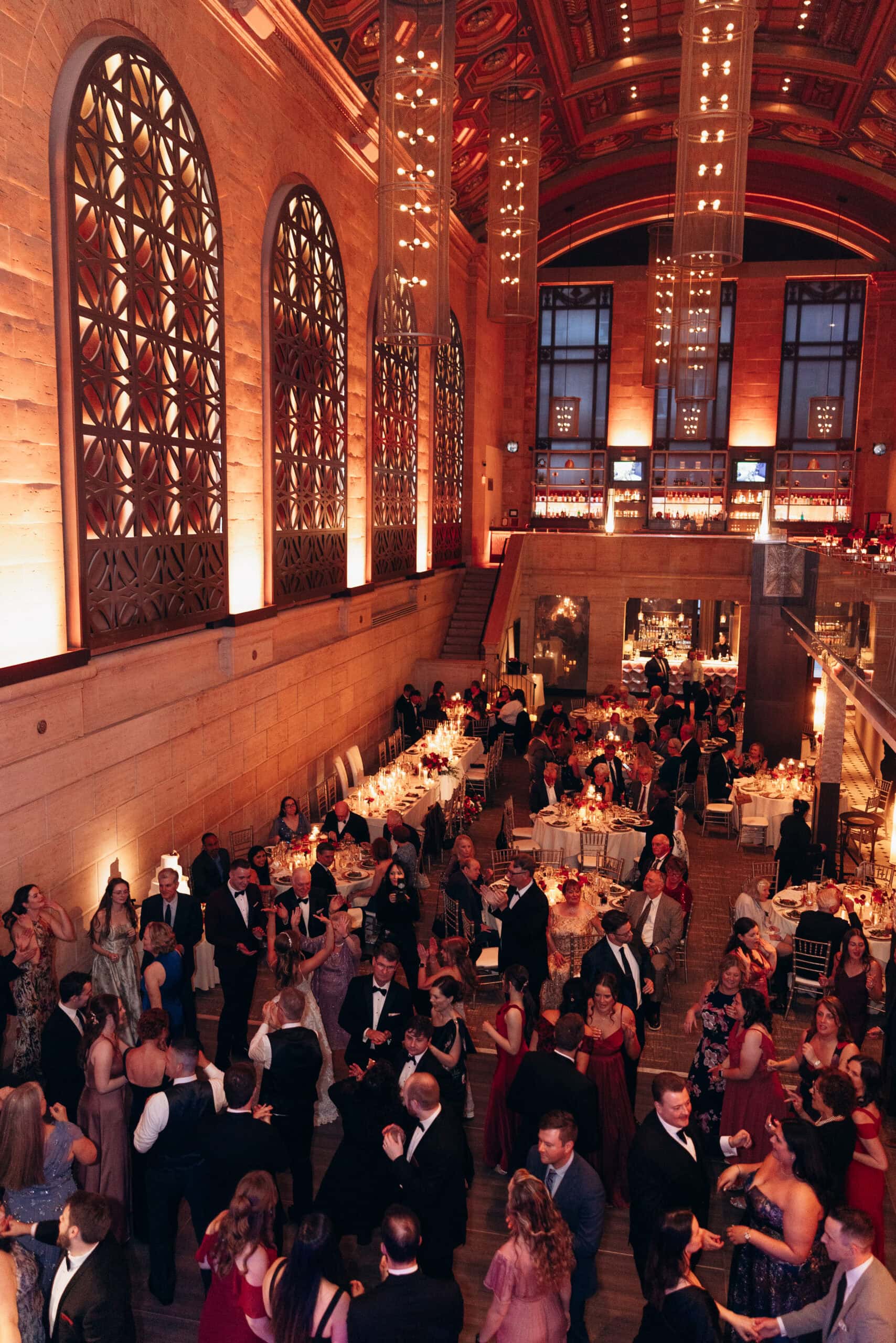 Interior of Union Trust Philadelphia with ornate windows, warm lighting, and guests in formal attire dining and dancing at a wedding reception.