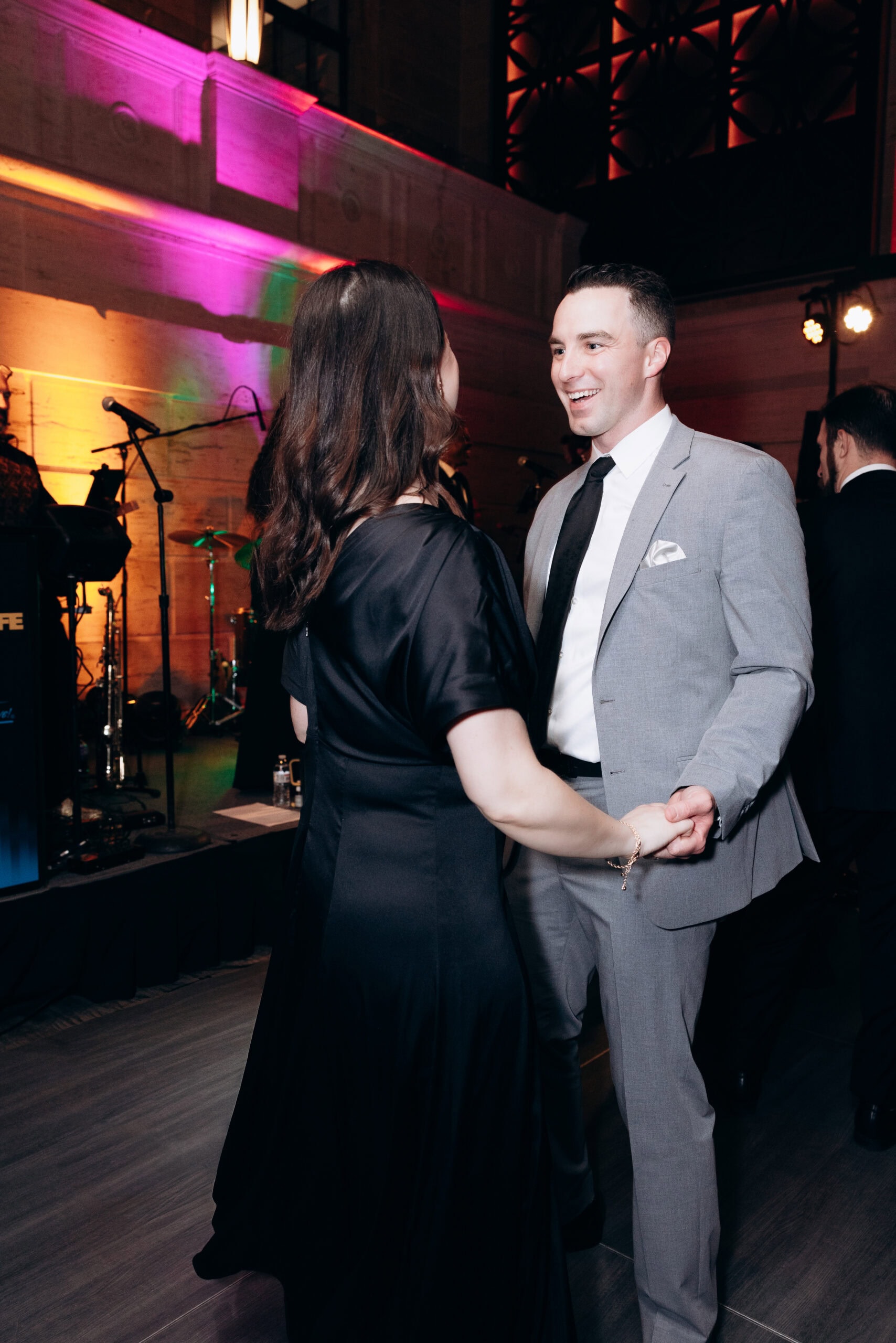 Man in light grey suit and black tie dancing with woman in black dress under colorful ambient lighting at Union Trust, Philadelphia.