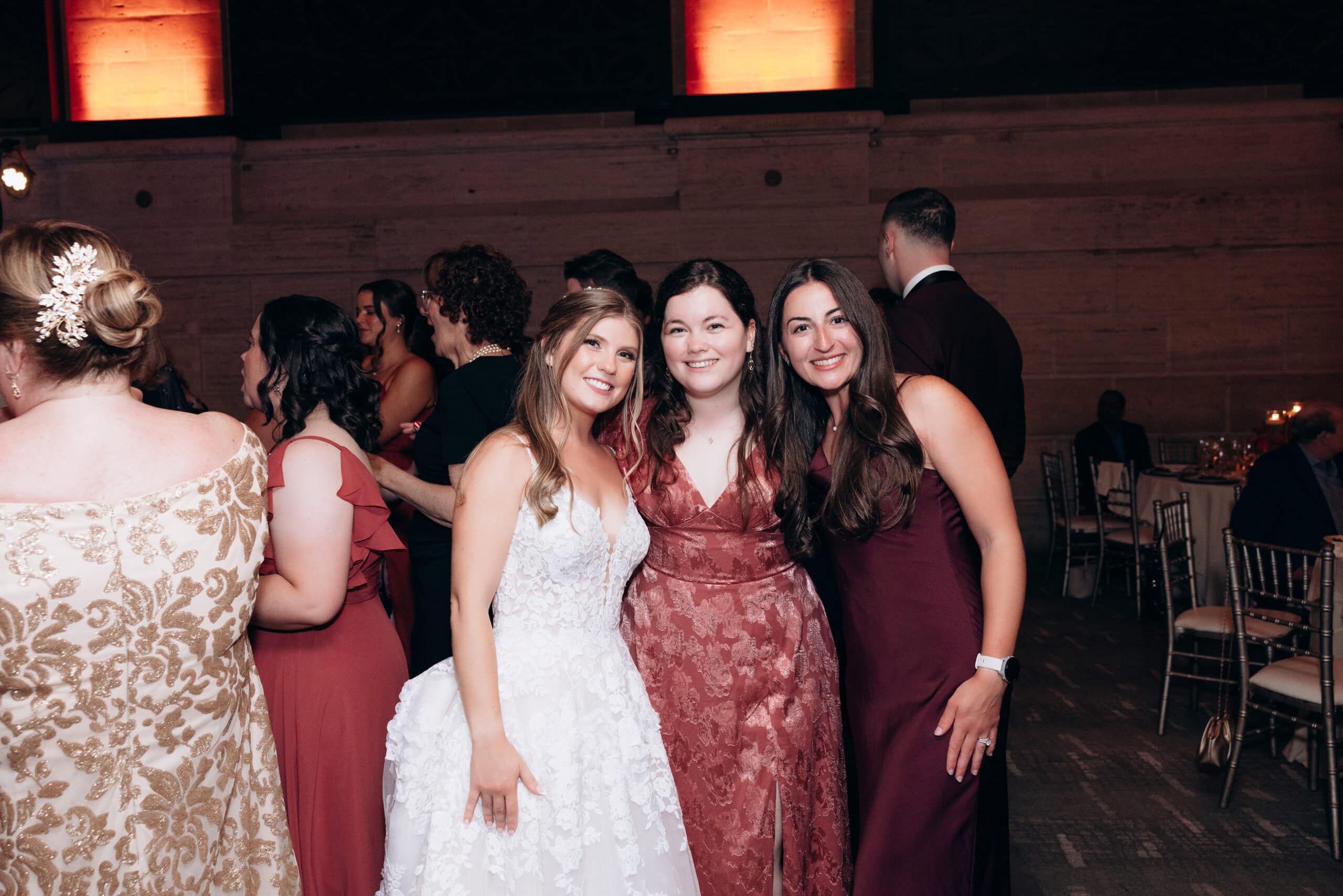 Bride in white lace gown posing with two women in deep red dresses at an indoor wedding reception with amber lighting and stone walls at Union Trust, Philadelphia.