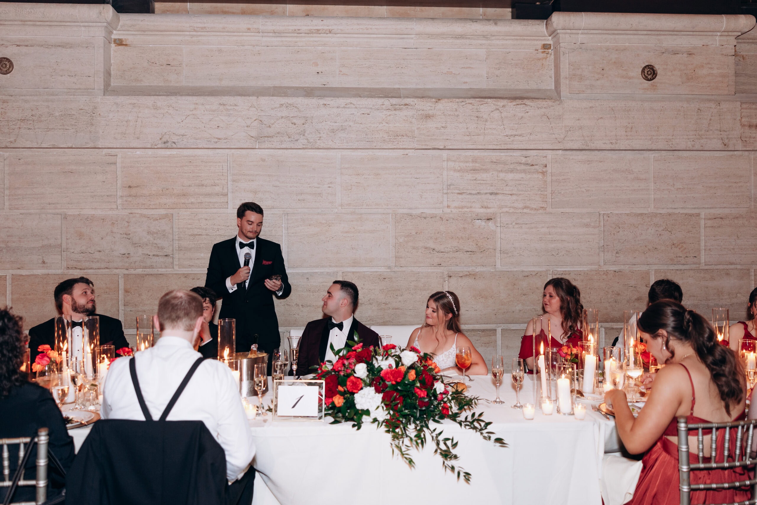 Man in tuxedo giving a speech with microphone at a wedding head table decorated with red and white flowers and candles, guests seated listening.