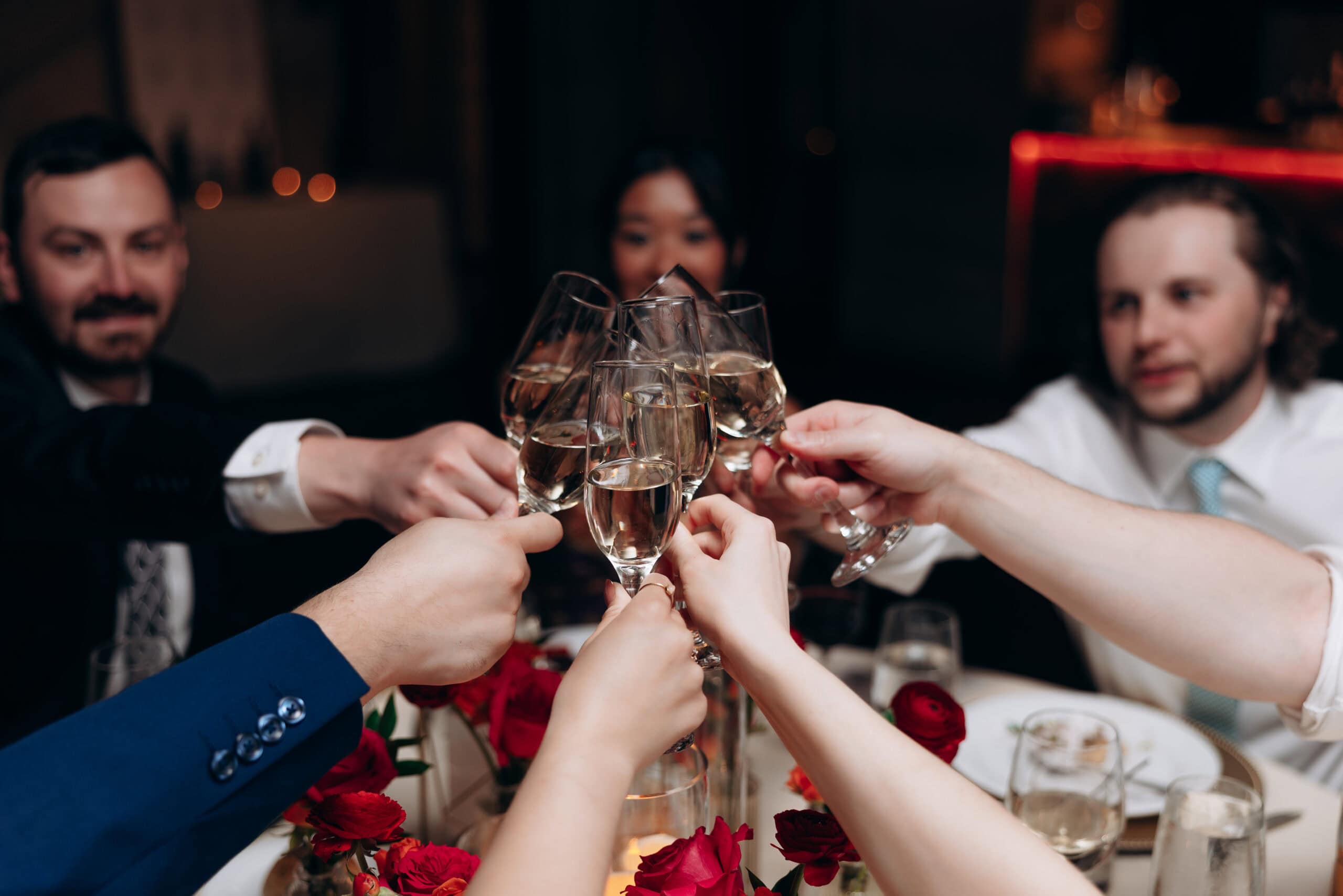 Close-up of multiple hands holding champagne flutes in a toast over a table decorated with red roses, with blurred guests in the background.