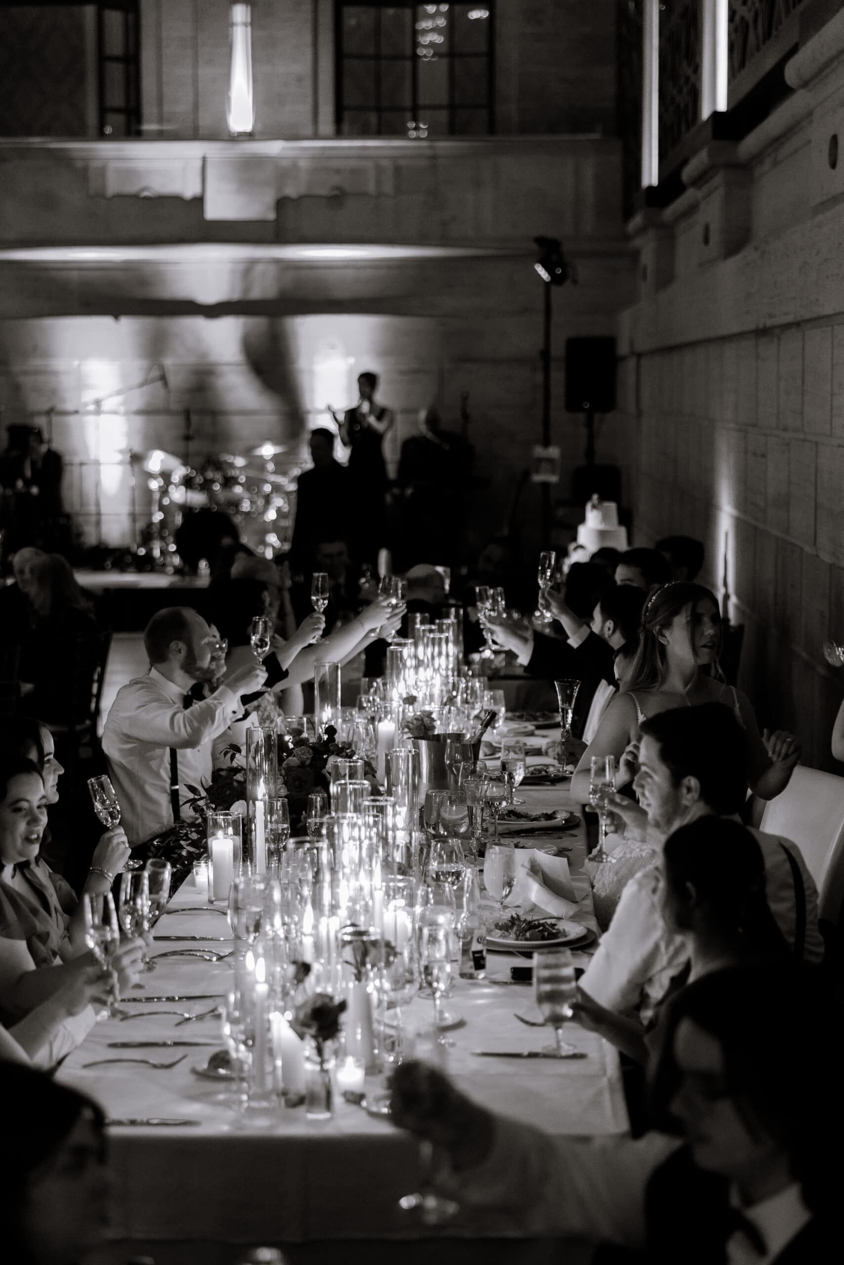 Guests seated at a long candlelit table raising glasses in a toast during a wedding reception at Union Trust, Philadelphia, with a live band in the background.
