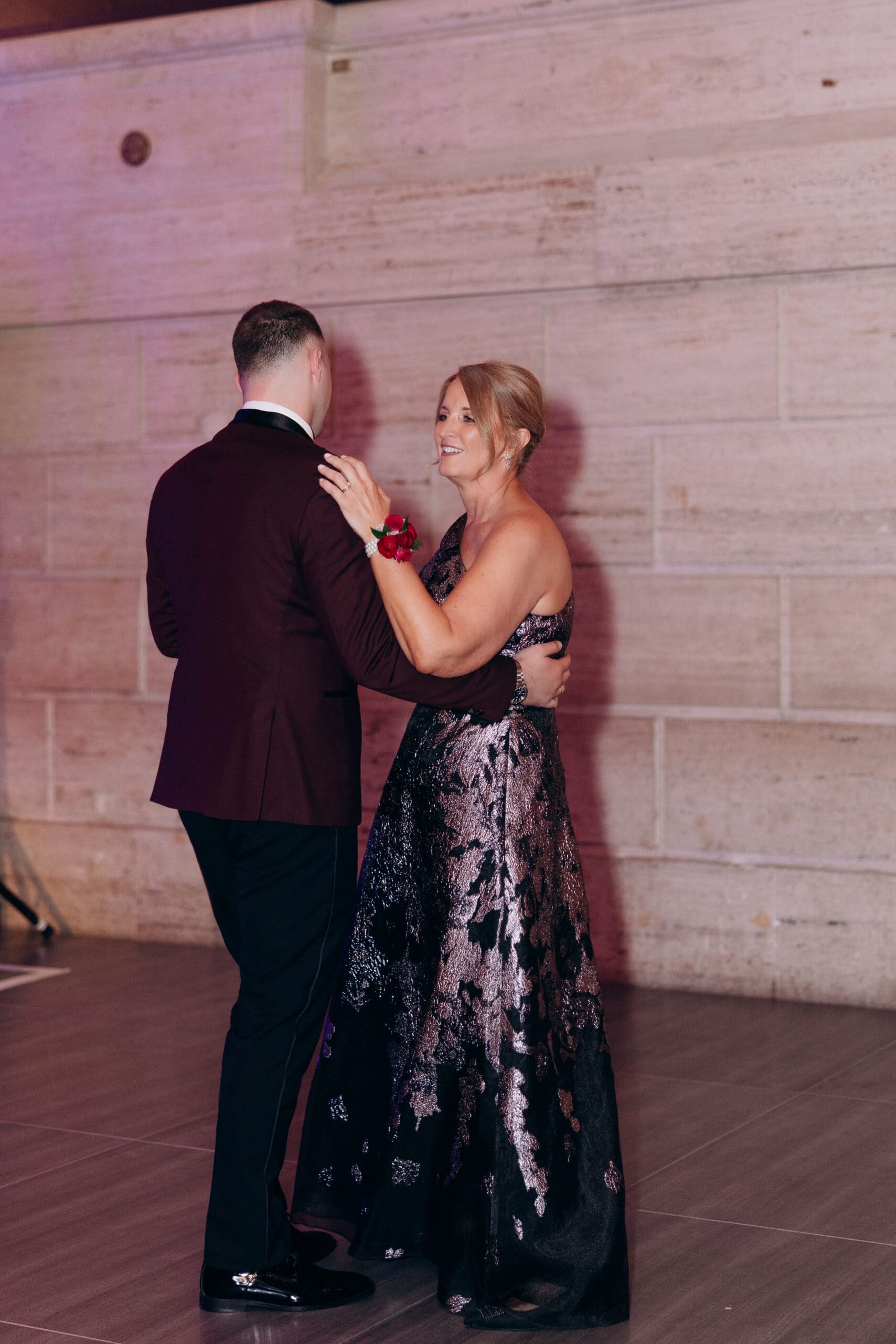 Mother and son dancing closely in formal attire at Union Trust Philadelphia, with stone wall background and soft lighting.