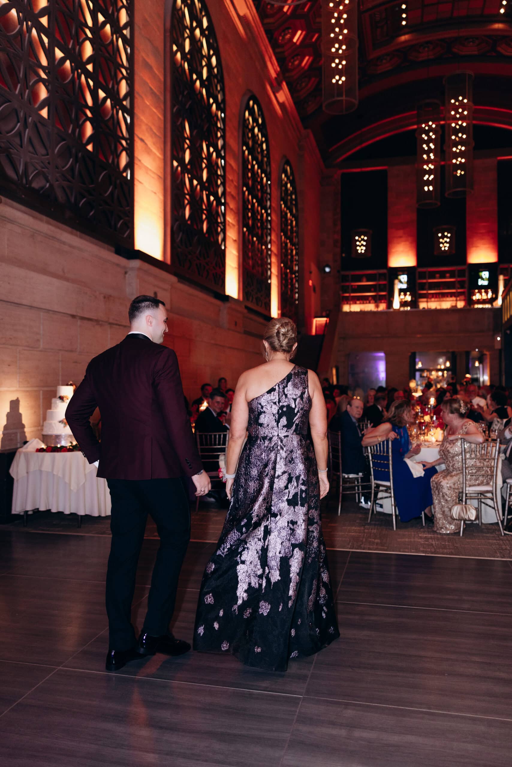 Couple entering a grand hall with ornate windows and warm lighting, guests seated at tables in the background at Union Trust, Philadelphia.