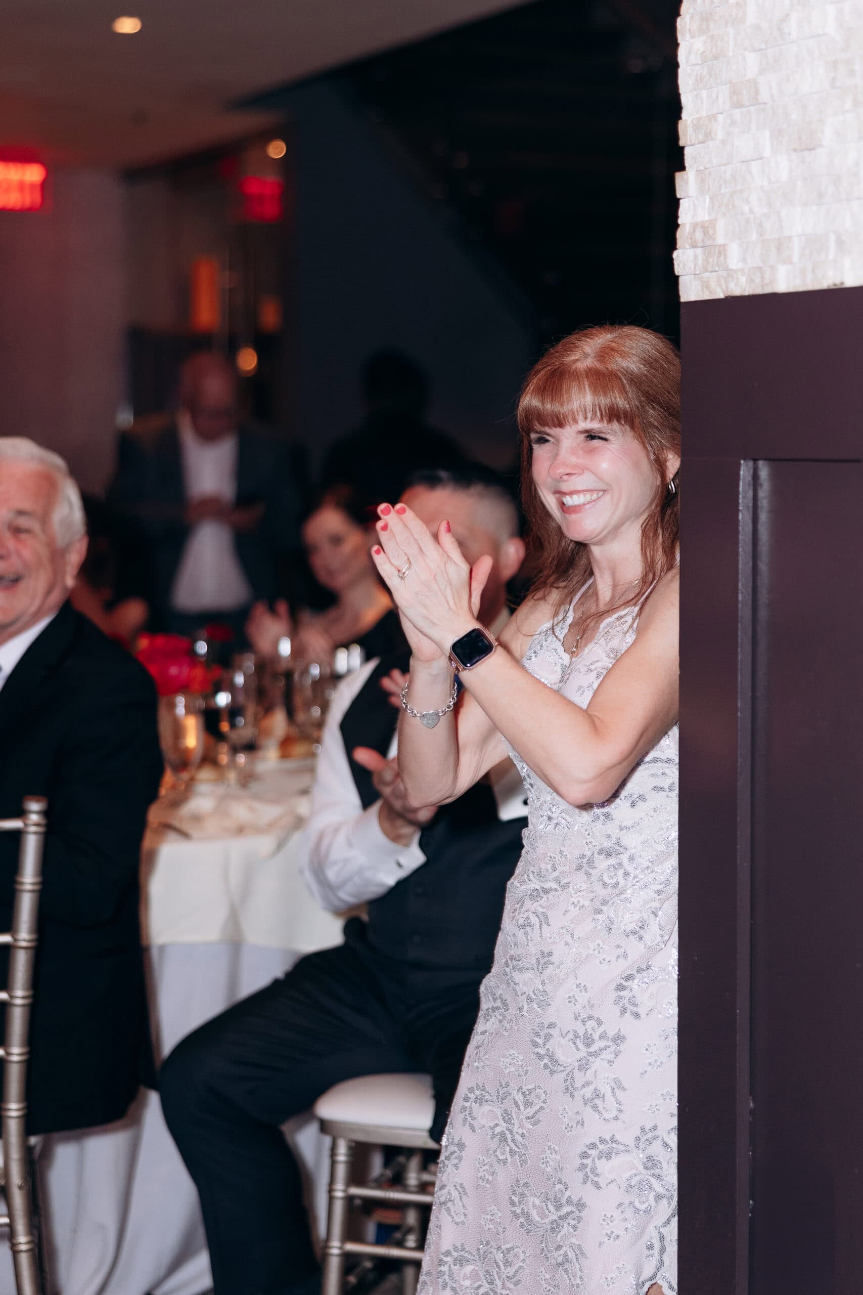 Woman in lace dress clapping and smiling during a wedding reception at Union Trust, Philadelphia, with guests seated at tables in the background.